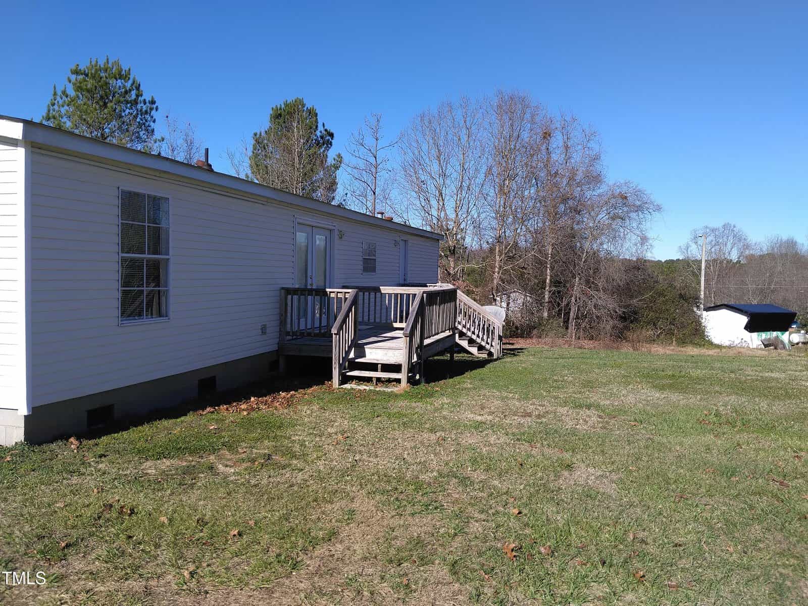 A single-wide mobile home with a wooden entrance deck sits on a grassy area under a clear blue sky, with trees in the background and a small boat covered with a tarp on the right side.
