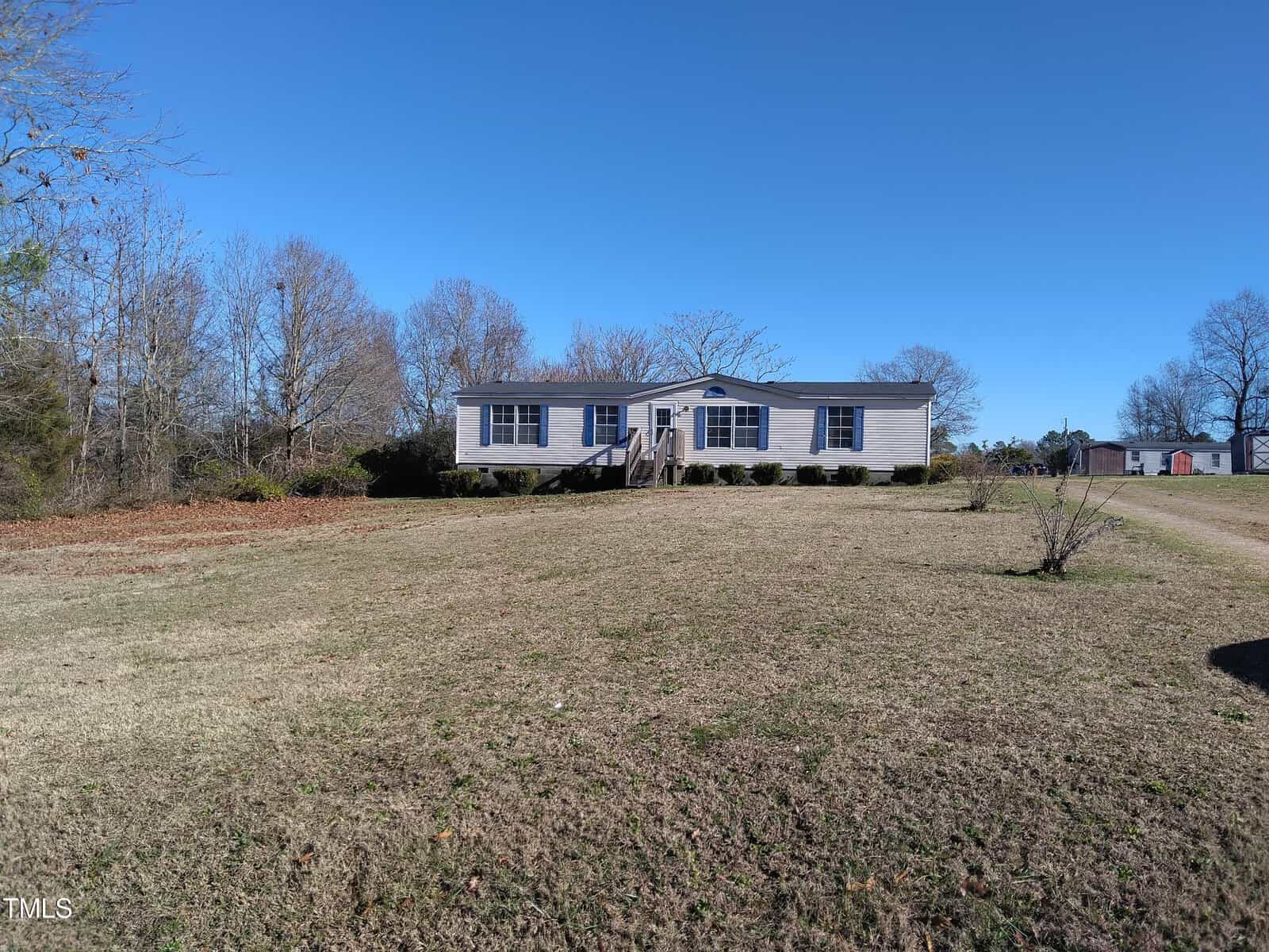 A single-story modular home with a central front entrance sits on a grassy plot with a clear blue sky overhead. trees flank the structure, and there are no leaves on the ground, suggesting it may be either late fall or winter. a driveway on the left appears to lead up to the home.