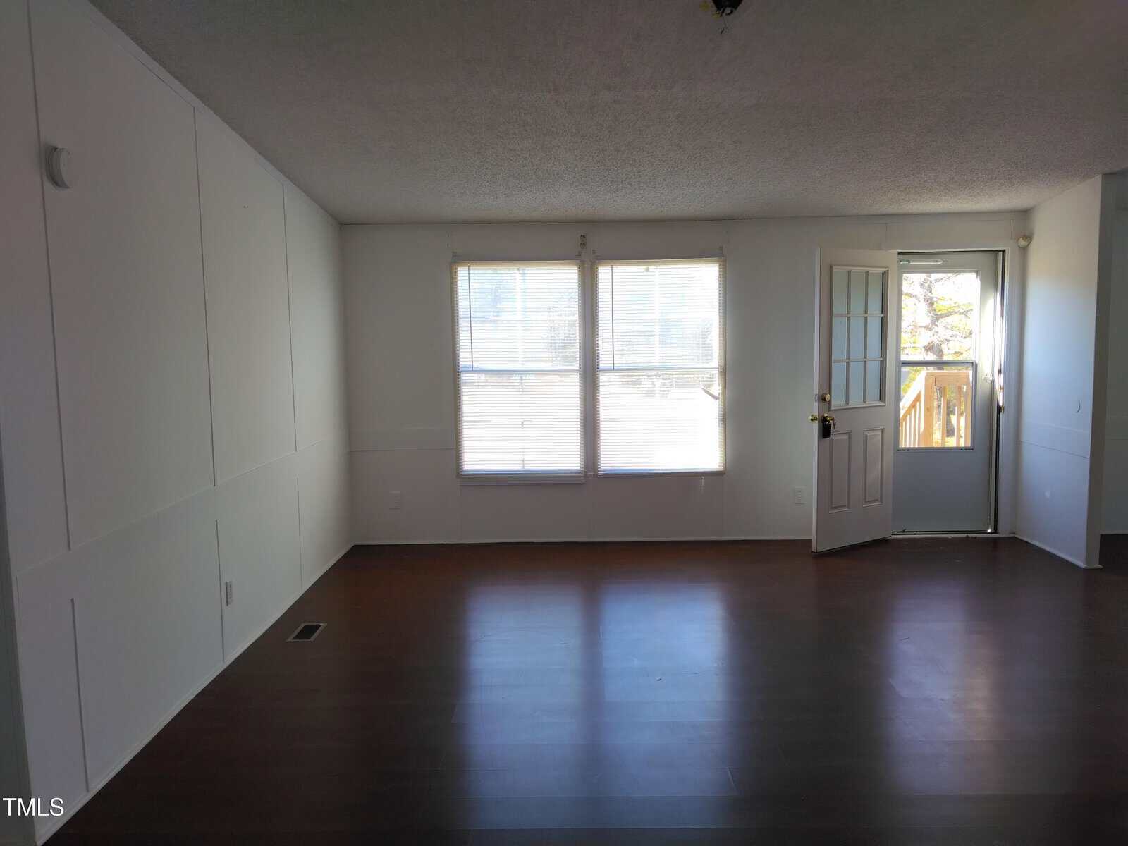 An empty room with hardwood floors, white walls, and a single window with blinds partially drawn near an open door leading to another room.
