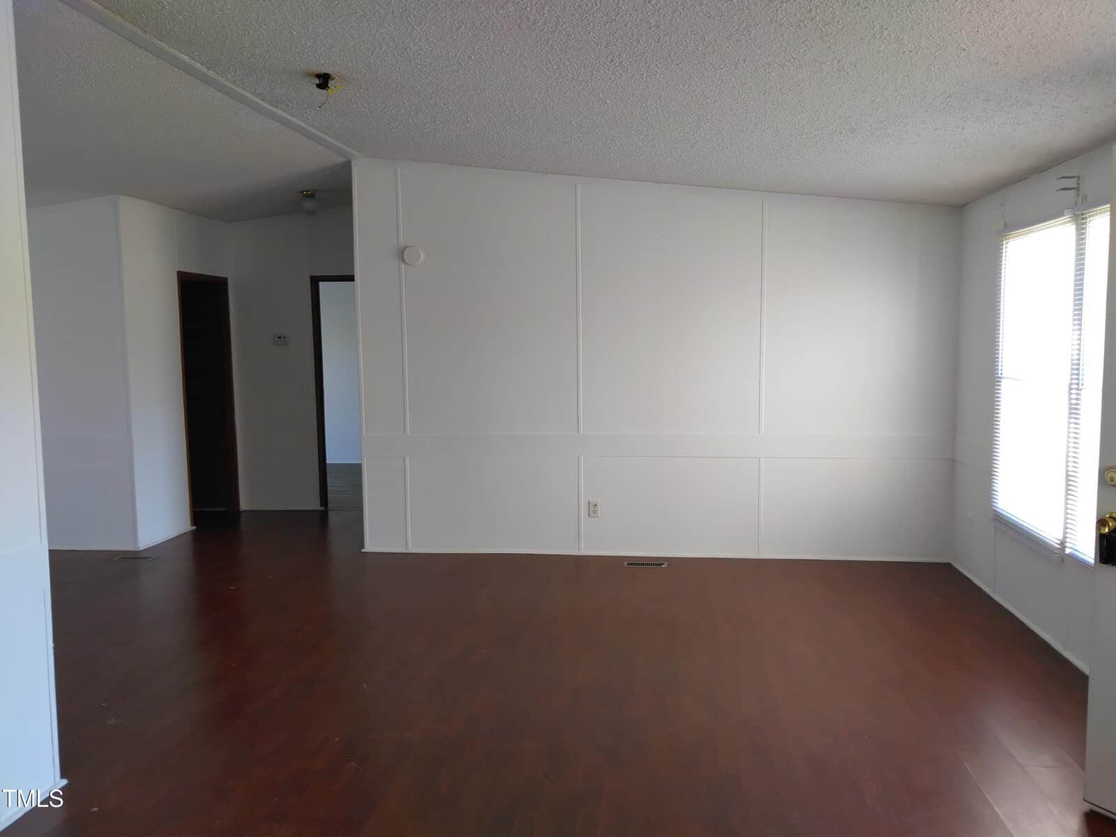 An empty room with white walls and a dark wooden floor, featuring a central heating vent on the floor and one window providing natural light.