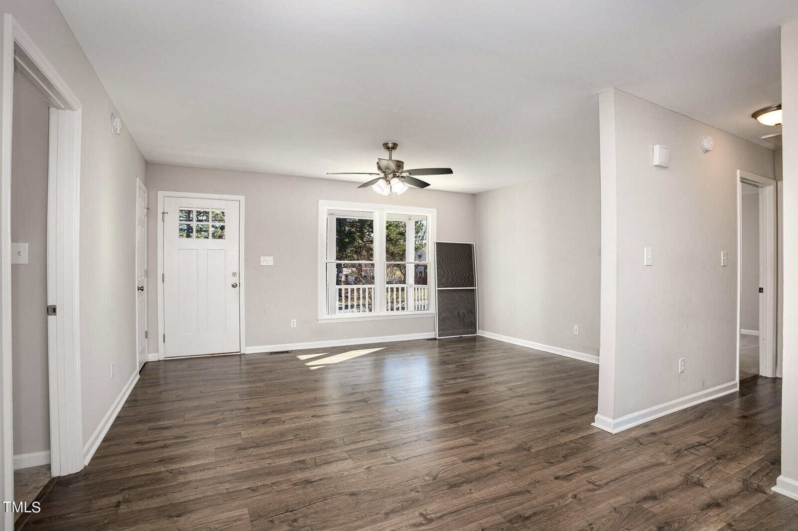 An empty, well-lit living room featuring shiny hardwood floors, white walls, a ceiling fan, and a glass-paneled door leading outside. a fireplace is visible along one wall.