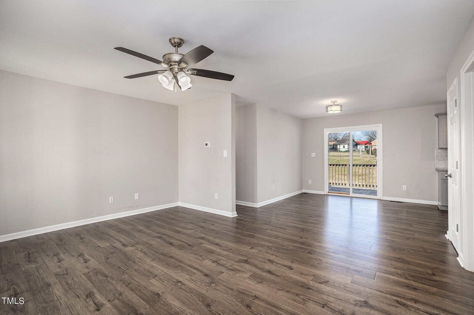Empty living room with hardwood floors, white walls, and a ceiling fan. large windows provide natural light, and a partially visible backyard can be seen.