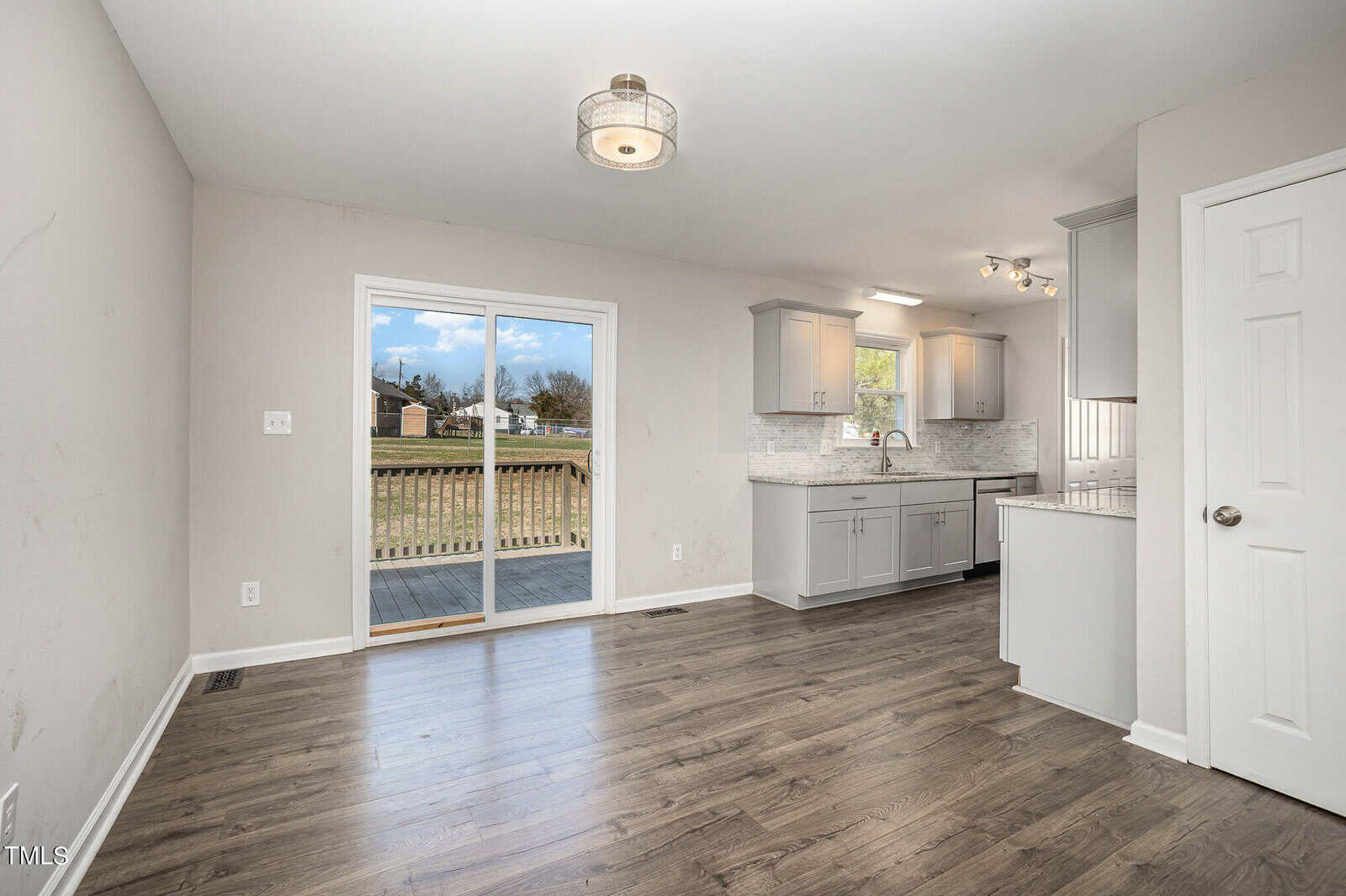 An empty room with gray hardwood floors, white walls, and a large glass sliding door leading to a backyard. it features a kitchenette on the right side with white cabinetry and modern appliances.