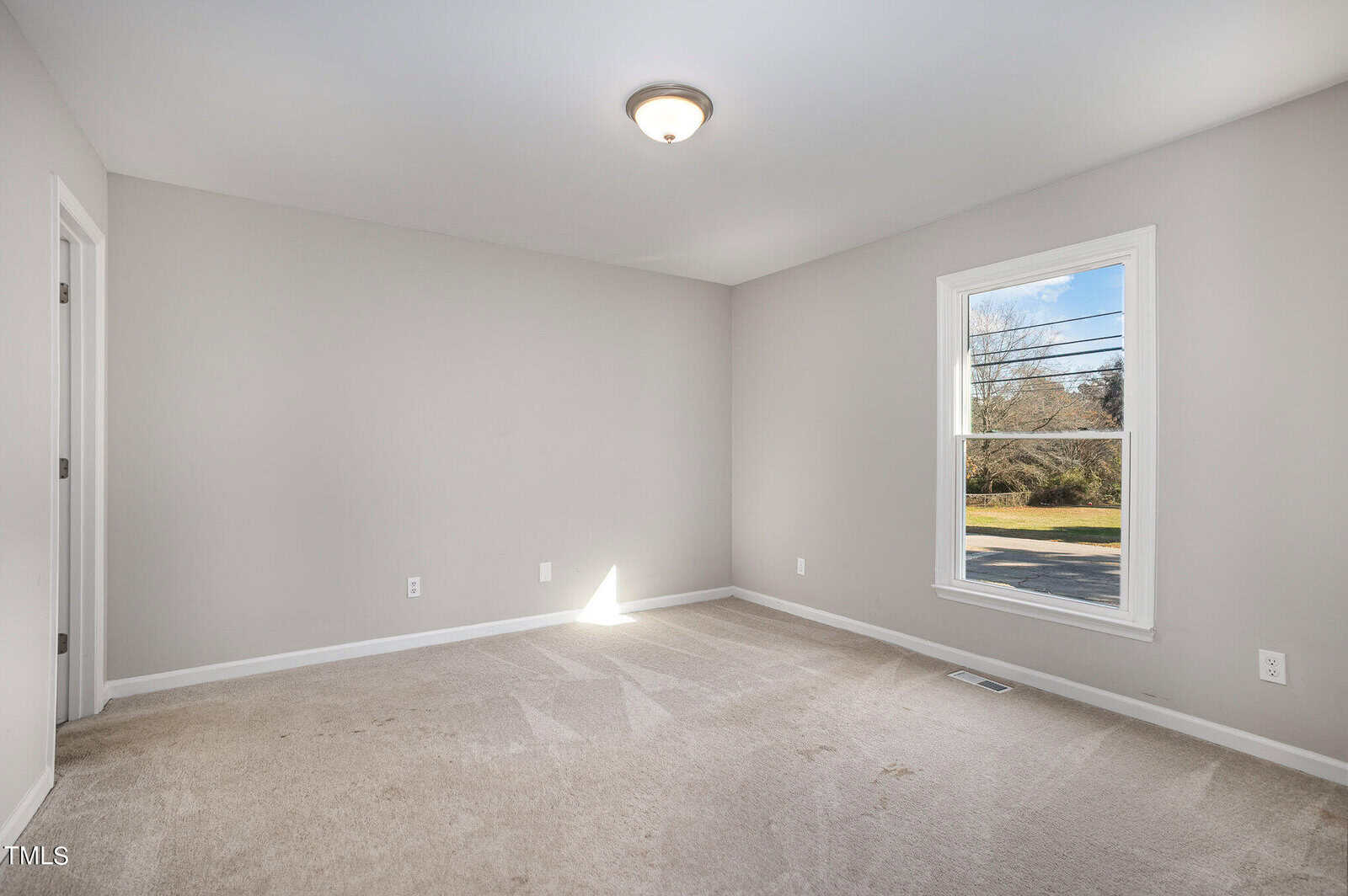 Interior of a vacant room with beige walls and carpeted flooring. features include a large window with a view of a street and a simple ceiling light fixture.