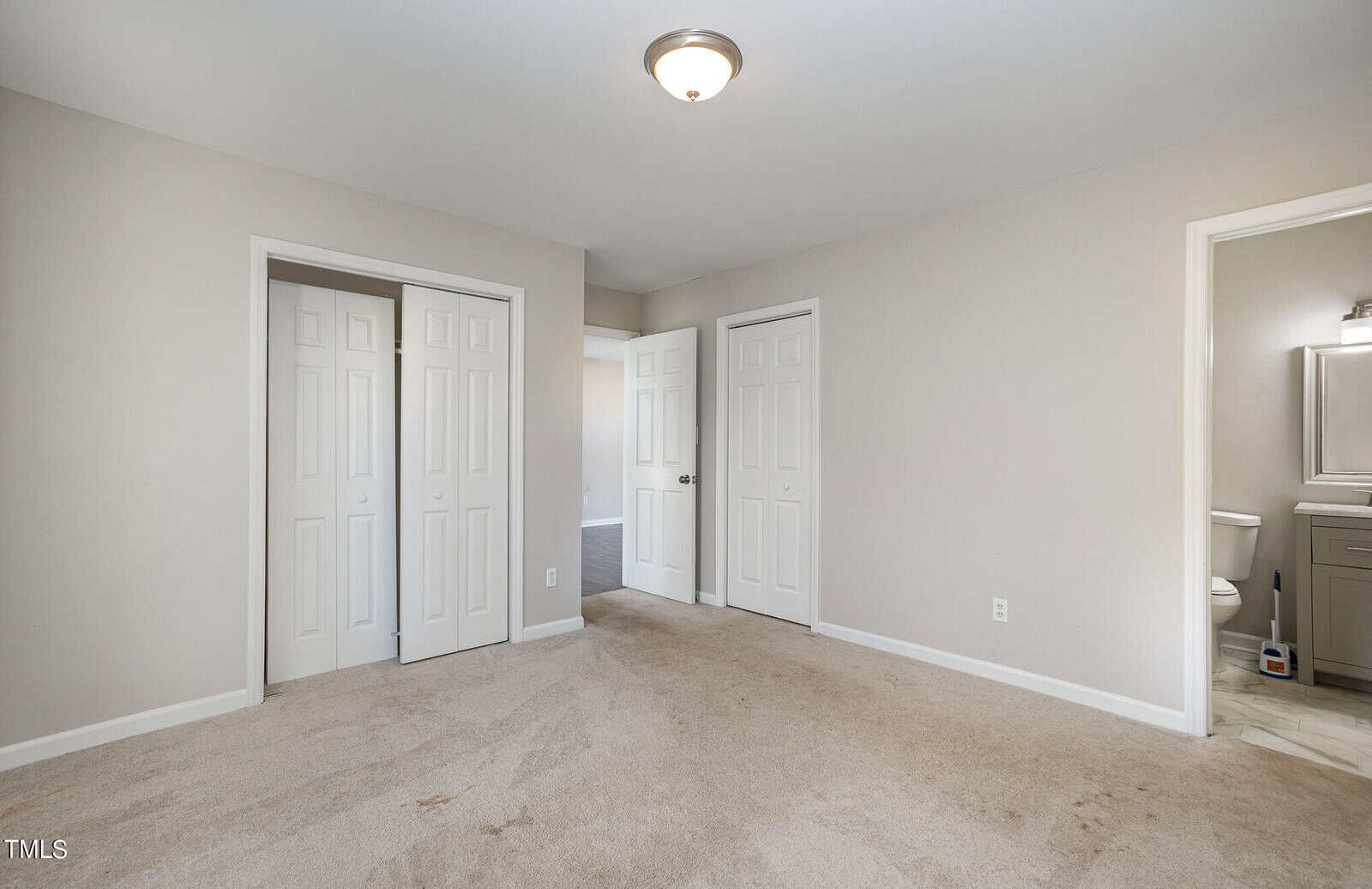 An empty room featuring a beige carpet with two white closed doors on one wall and an open door leading to a bathroom on the adjacent wall. a small ceiling light illuminates the space.