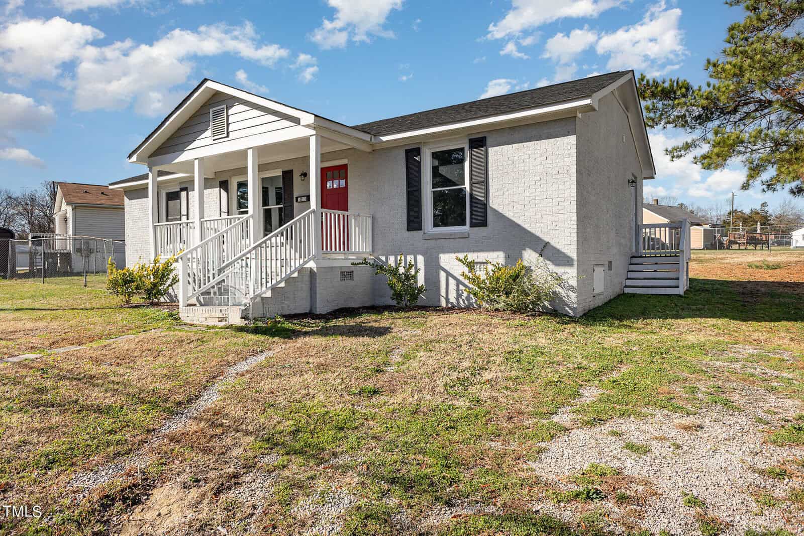 A small, single-story white house with a red door, located at 1013 N Pinkston Street, Henderson NC. It features a sloping front yard and a gravel path. The house has