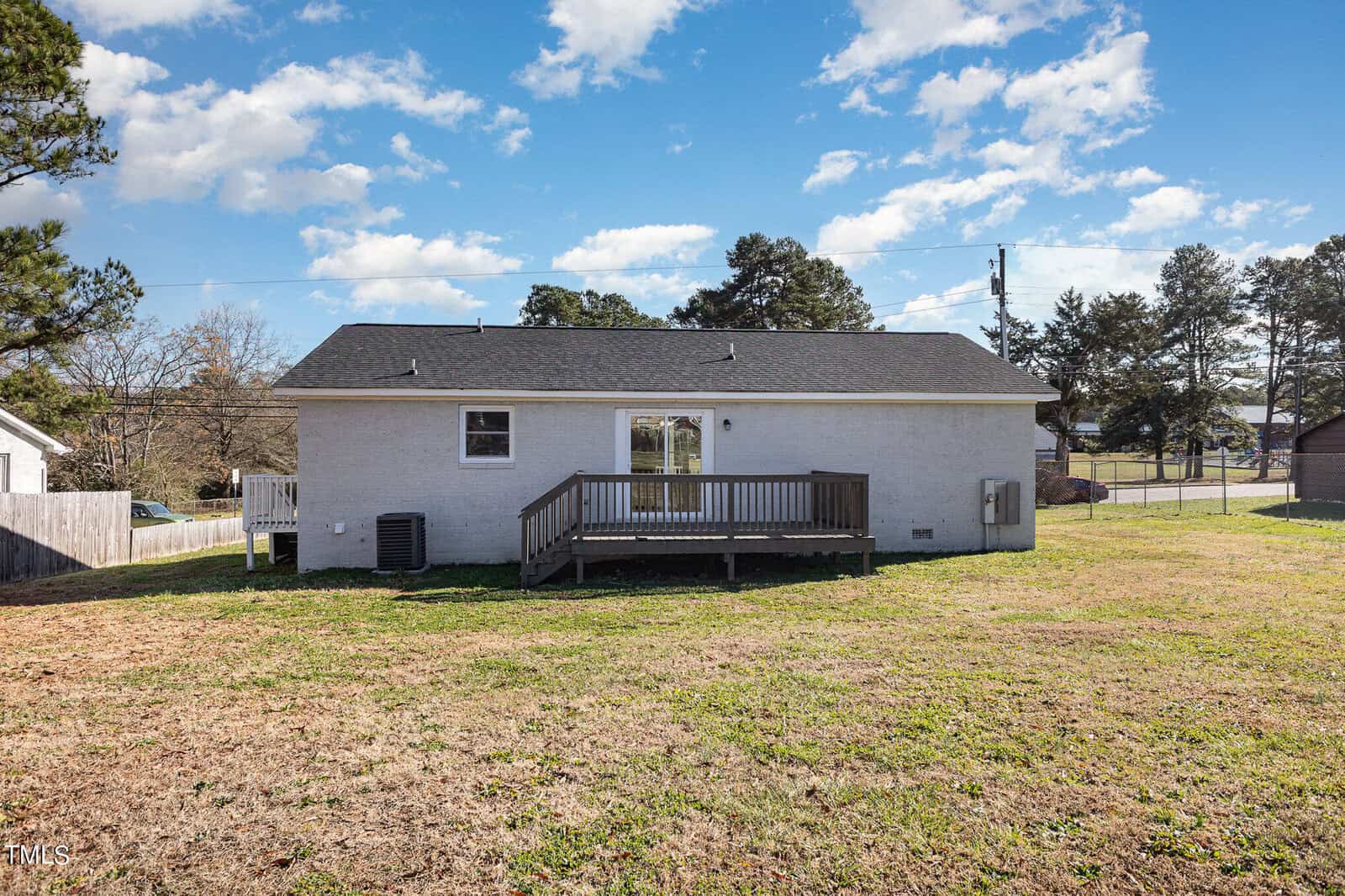 A small, white single-story home with a wooden deck and a grassy backyard, surrounded by a sparse neighborhood under a clear blue sky.