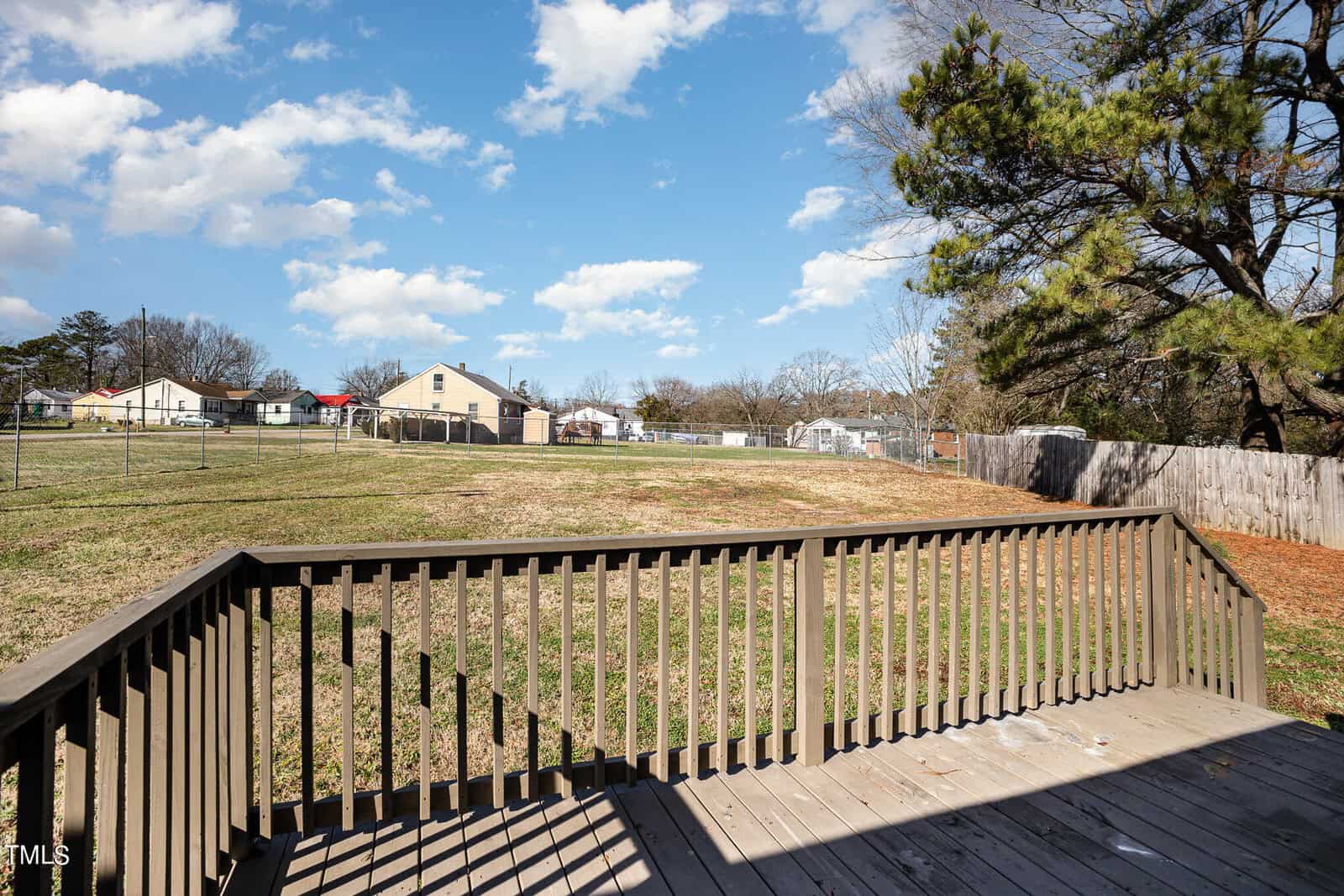 View from a wooden deck overlooking a spacious backyard with grass and a wooden fence. houses and trees are visible in the background under a clear blue sky.