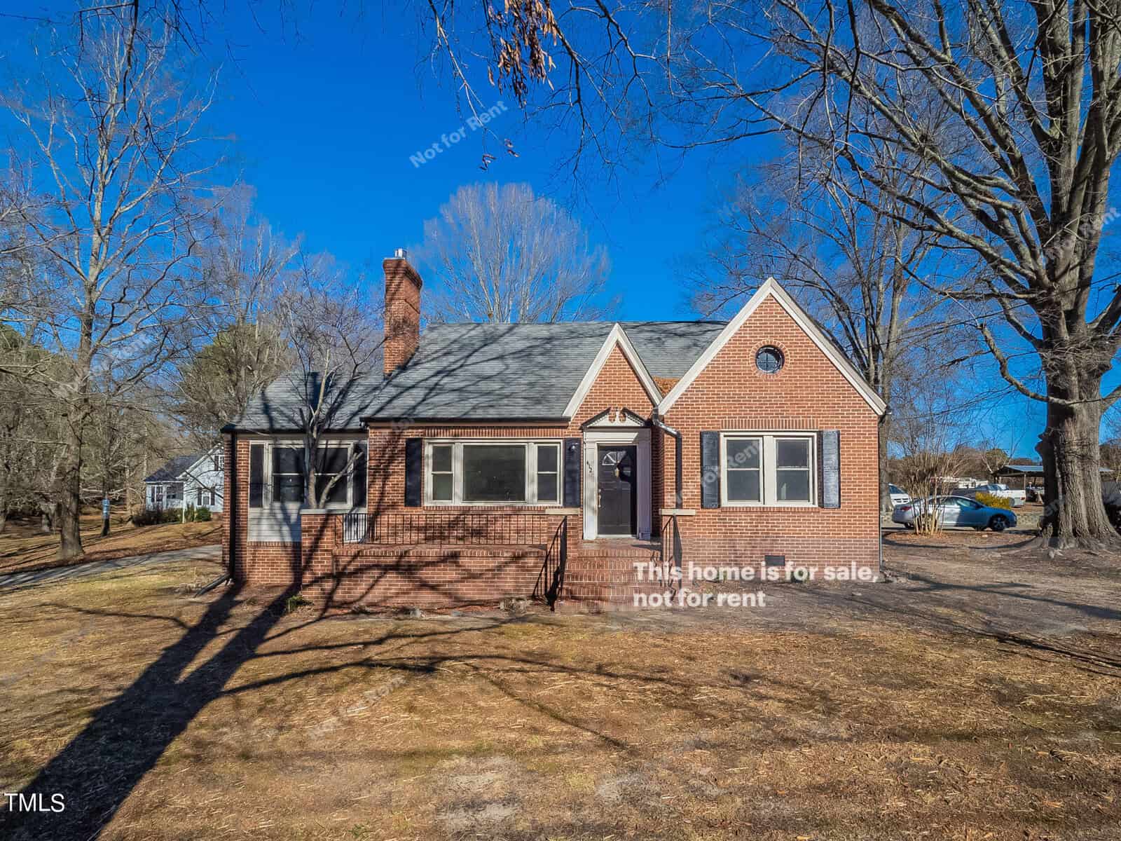 A brick house at 412 Lake Road, featuring a charming chimney and triangular roof, boasts a front entrance flanked by large windows. Leafless trees stand in the Creedmoor yard under a clear blue sky, while the ground is scattered with dry leaves and grass.