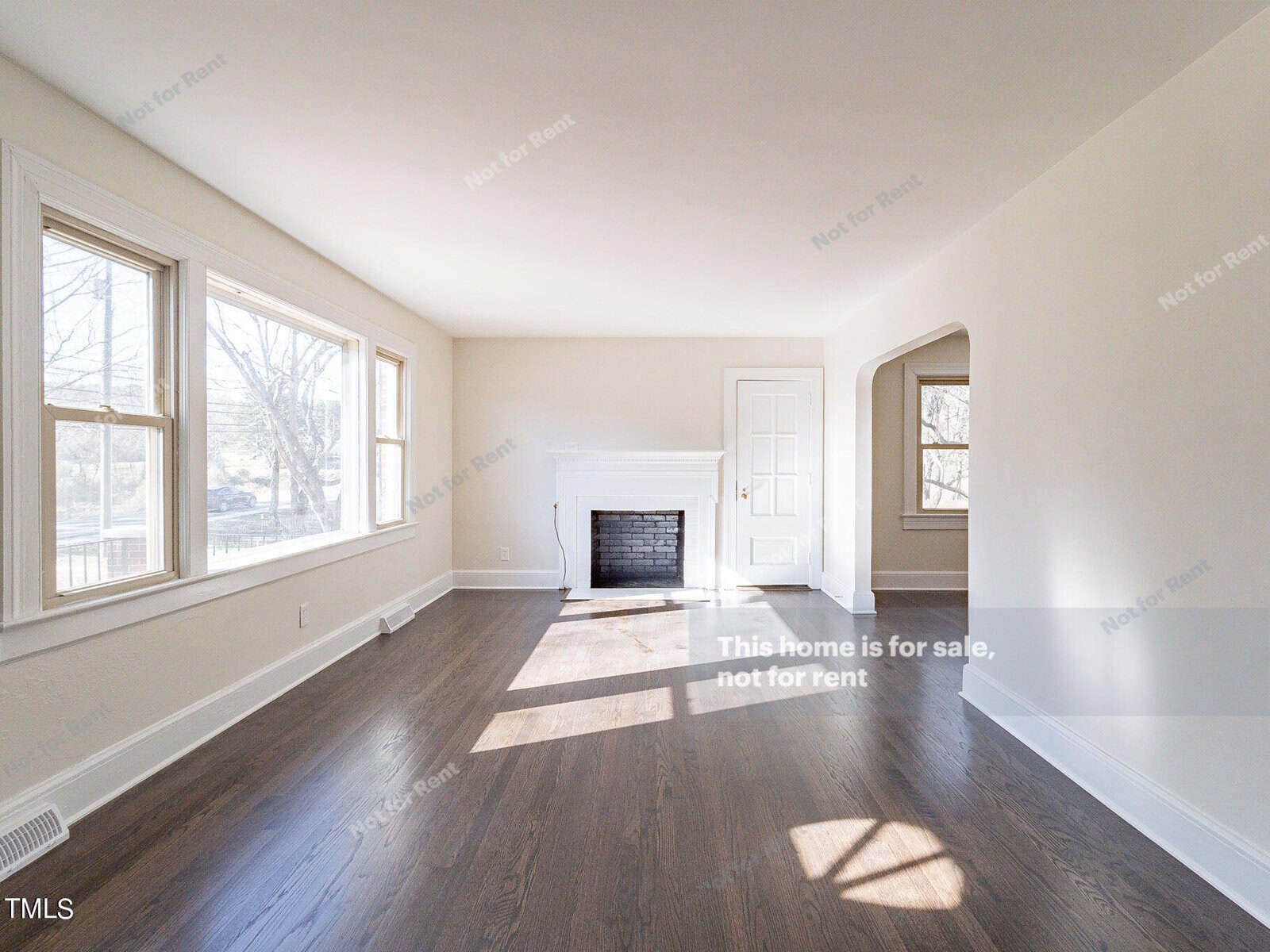 A bright, empty living room with hardwood floors, white walls, a fireplace, large windows, and a closed door. Sunlight streams through the windows, casting shadows on the floor. The room is unfurnished and bare.
