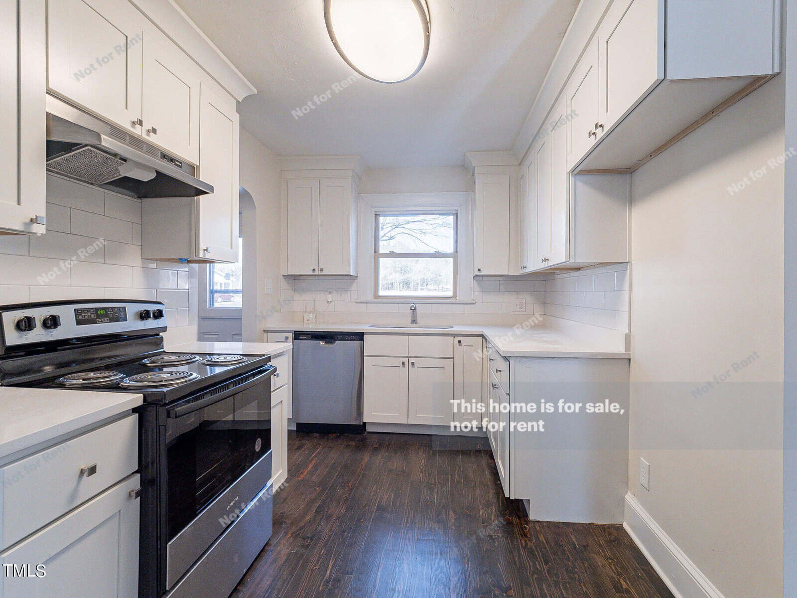 A modern kitchen with white cabinets, a stainless steel oven, dishwasher, and a double sink. The room has dark hardwood floors, a large central light fixture, and a window above the sink. Text indicates the home is not for sale or rent.