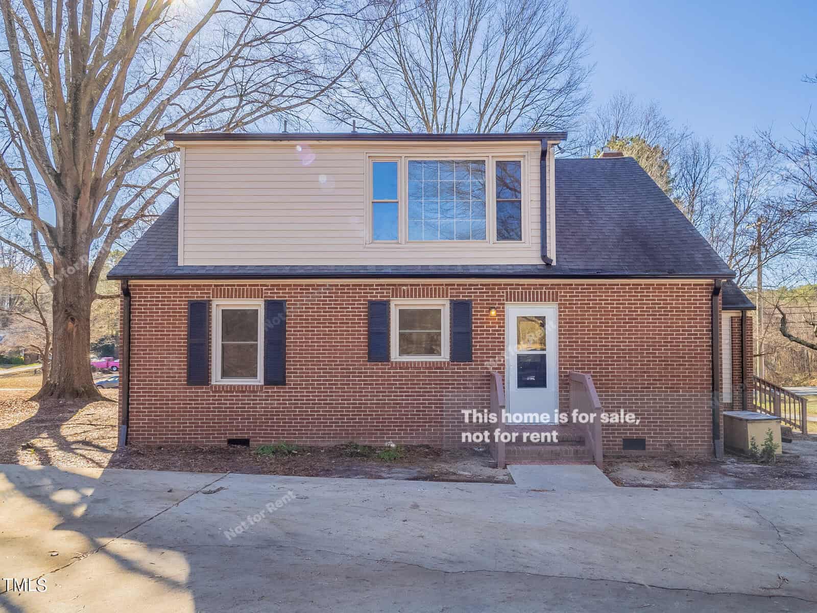 A two-story brick house with a sloped roof is situated in a natural setting with large trees. The first floor features a few windows with shutters, and a dormer window is present on the second floor. The driveway is visible in the foreground.