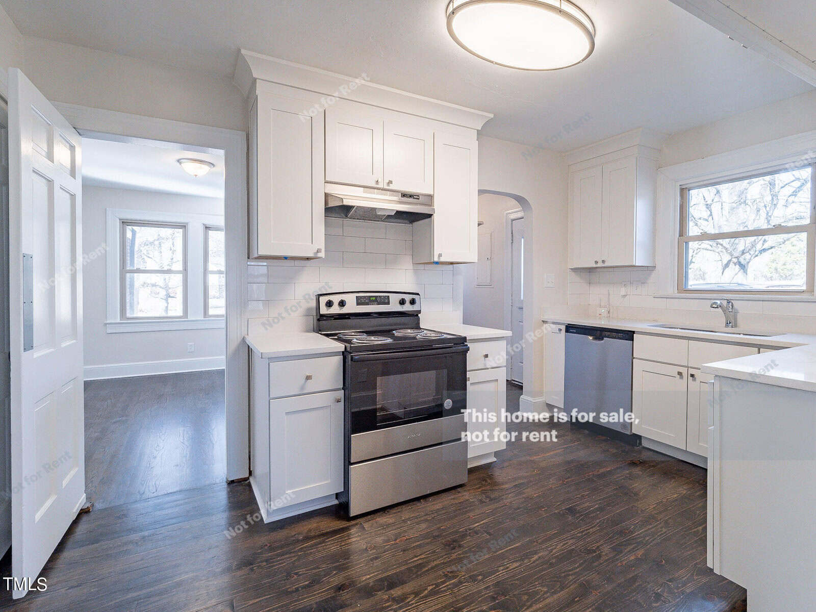 A modern kitchen with white cabinets, stainless steel appliances, and dark wood flooring. The room is well-lit with natural light from windows and features a round ceiling light. Text on the image states: "This home is for sale, not for rent.