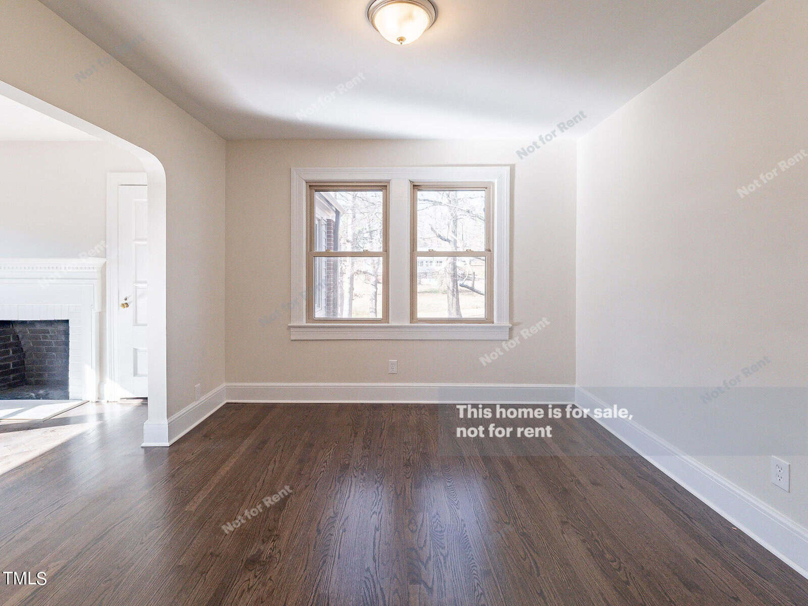 A bright, empty room with polished hardwood floors and a small ceiling light. There are two large windows, and the room connects to another area with a white fireplace. Text on the image reads, "This home is for sale, not for rent.