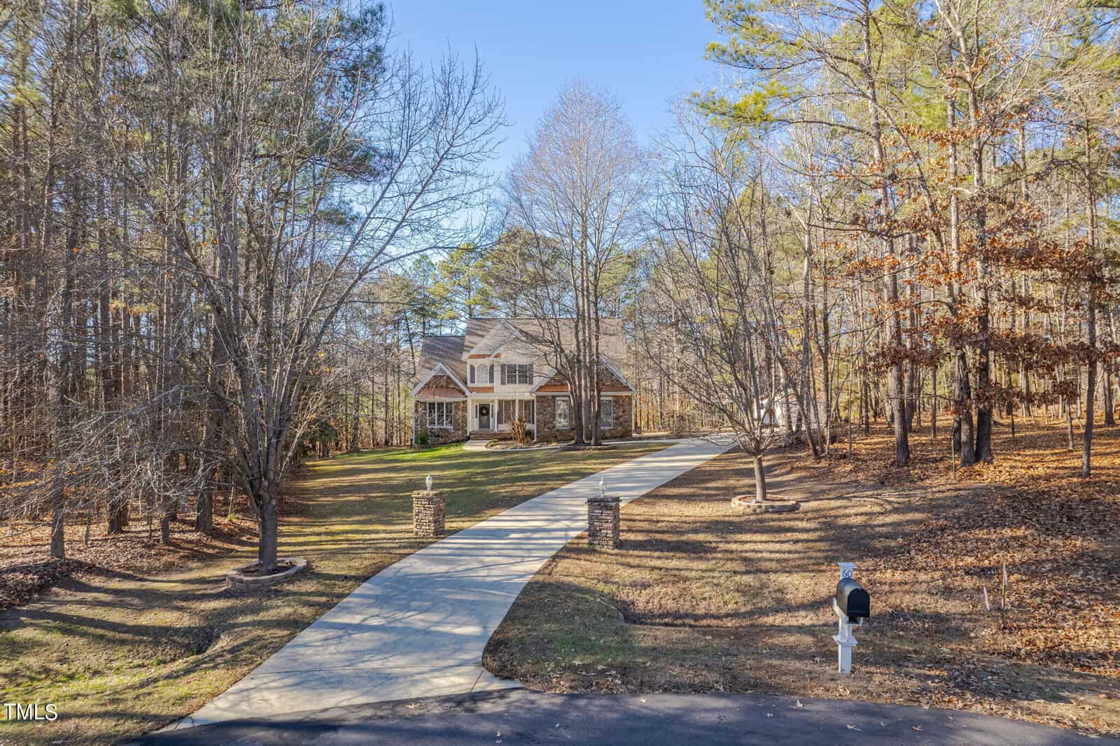 A large house with a gabled roof is nestled among bare trees. A long, curved driveway leads to the entrance, and a mailbox is visible near the road. The surrounding yard is spacious and covered with fallen leaves.