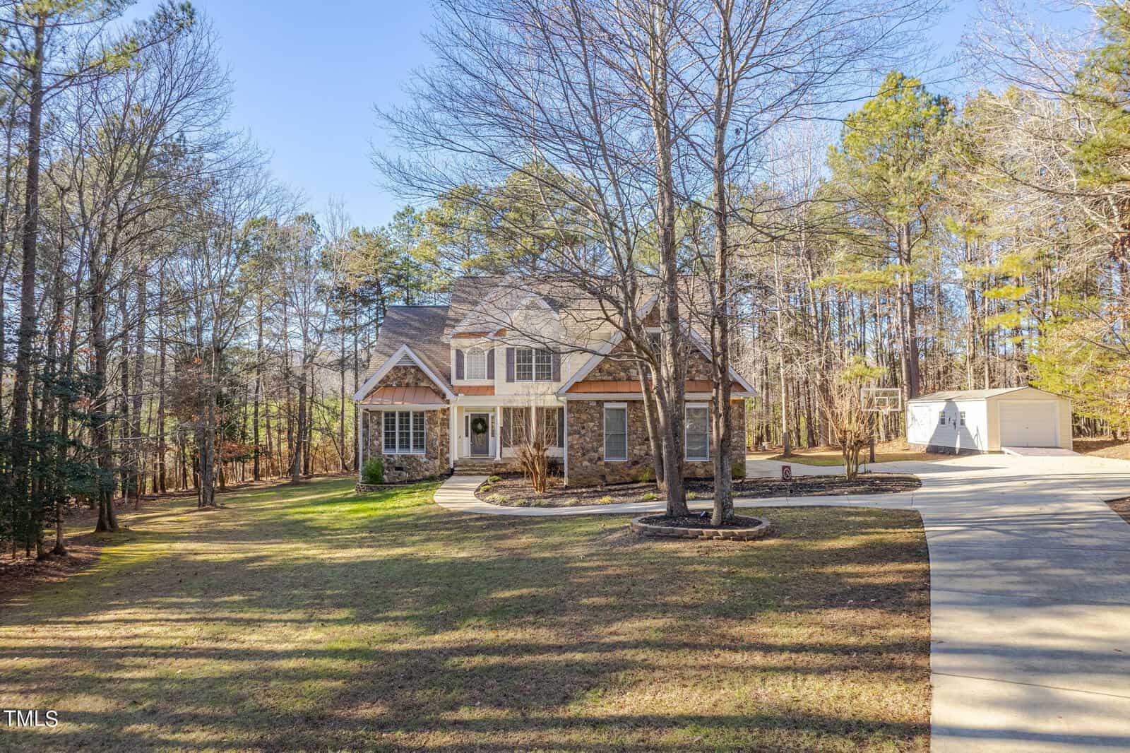 Two-story house with a gabled roof, surrounded by trees. A curving driveway leads to a detached garage on the right. The lawn is partially shaded by leafless trees, and the sky is clear and blue.