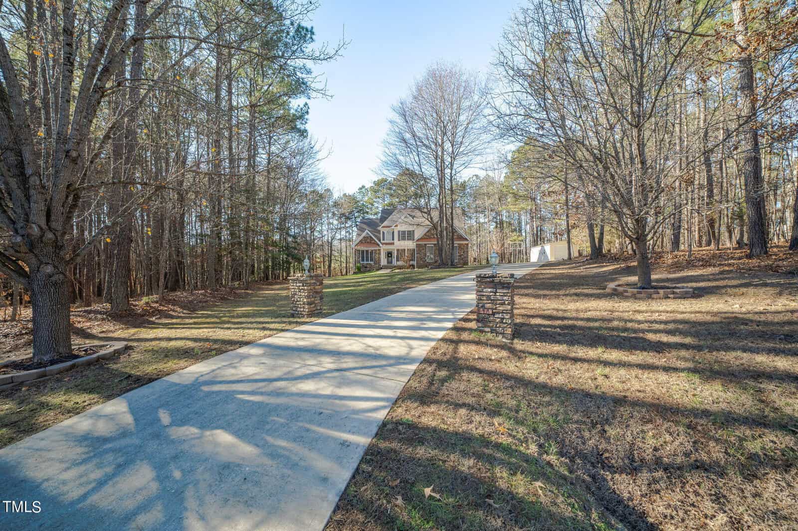 A curved driveway leads to a large house surrounded by tall trees. The pathway is bordered by two stone pillars, and the ground is covered with dry, wintry grass. The house is set against a backdrop of a clear blue sky.