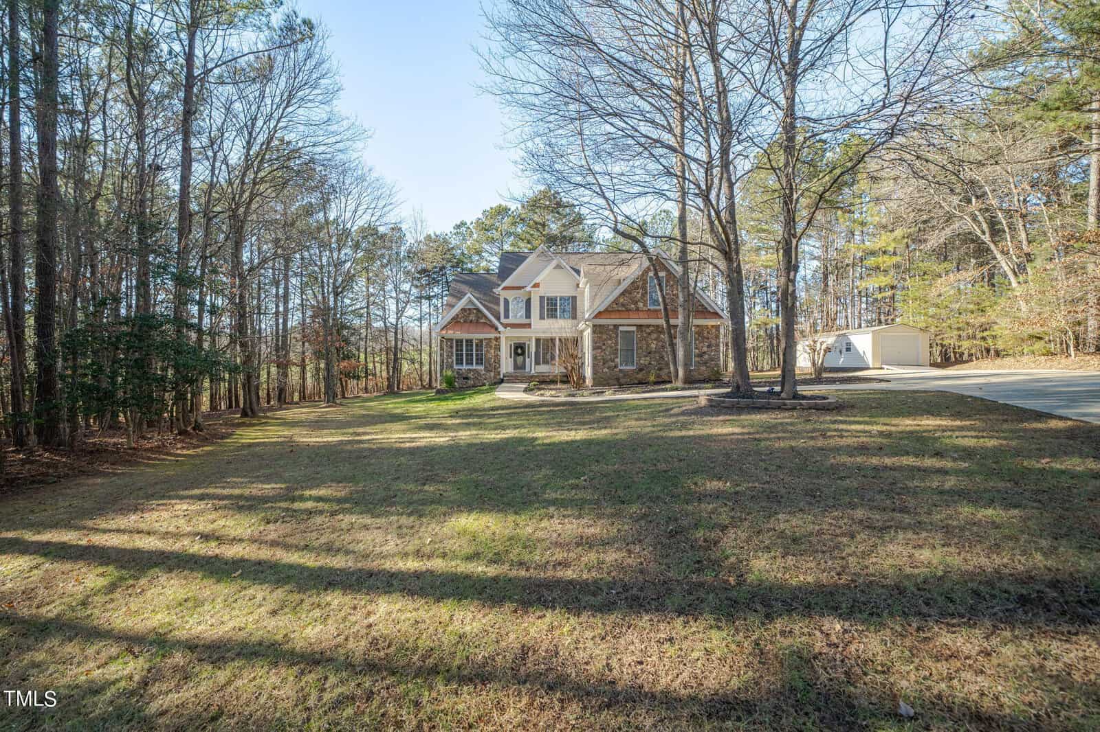 A two-story house with a front porch is surrounded by trees and situated on a spacious grassy lawn. A driveway leads to a detached garage. The area is wooded with tall trees and the sun casts shadows on the ground.