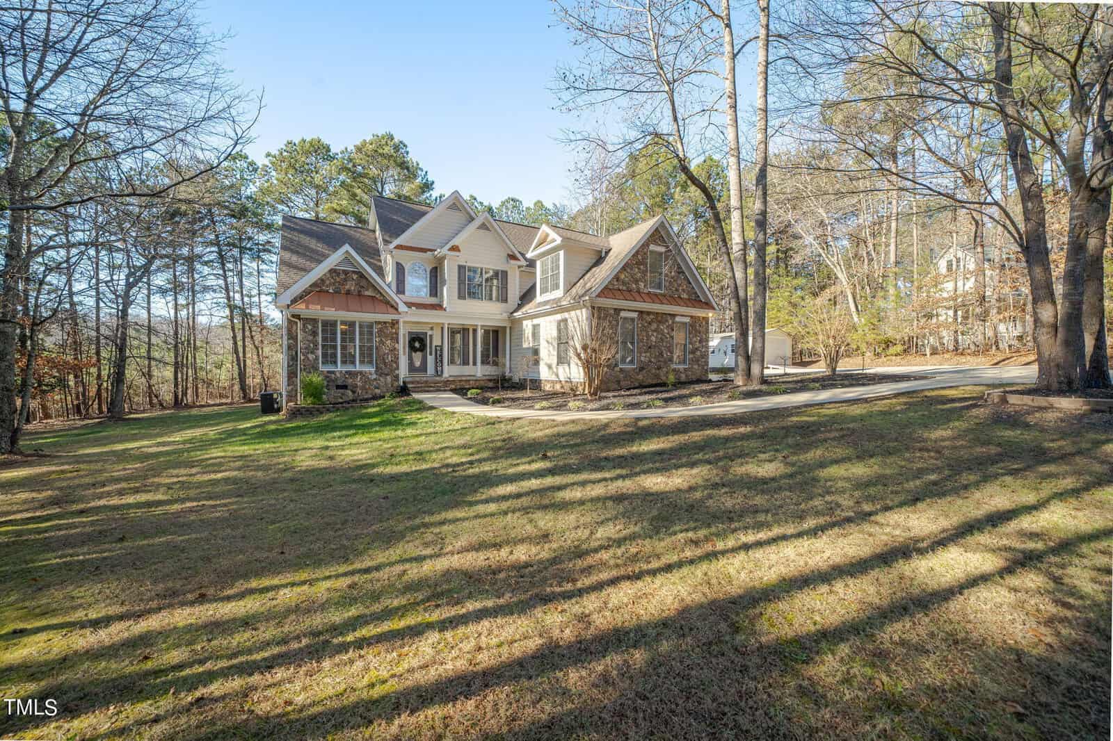 A large two-story house with stone and siding exterior surrounded by trees. It features a long driveway and a spacious front yard with grass. The sky is clear, indicating a sunny day.