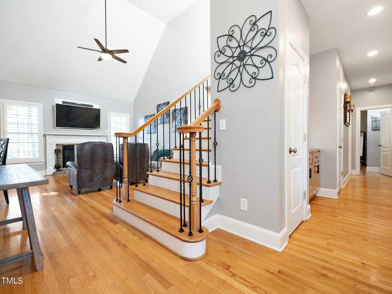A bright living room with wooden flooring and a staircase. The room features a fireplace, a TV above the mantel, and leather seating. Wall decor and a dining table are visible, with natural light streaming through large windows.