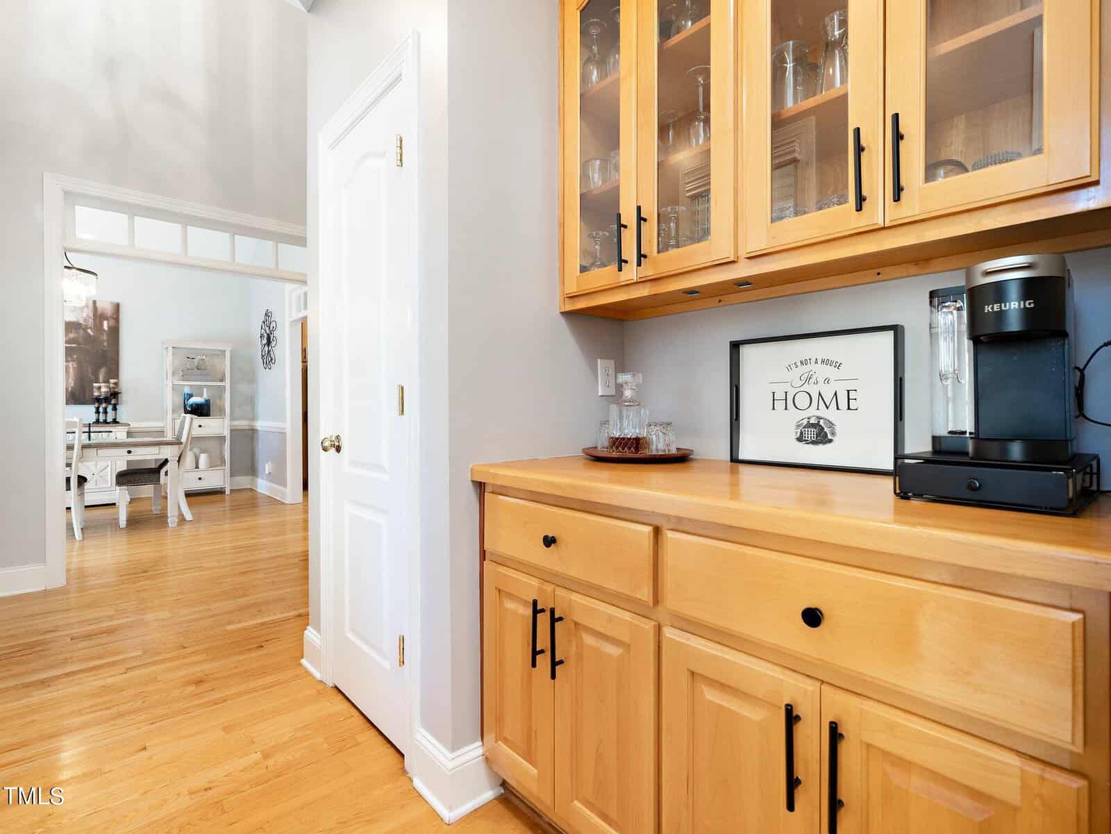 A cozy kitchen corner with a wooden cabinet featuring glass doors. The countertop holds a Keurig coffee maker and a sign saying "Home." Hardwood floors extend into a dining area visible in the background with a table and chairs.