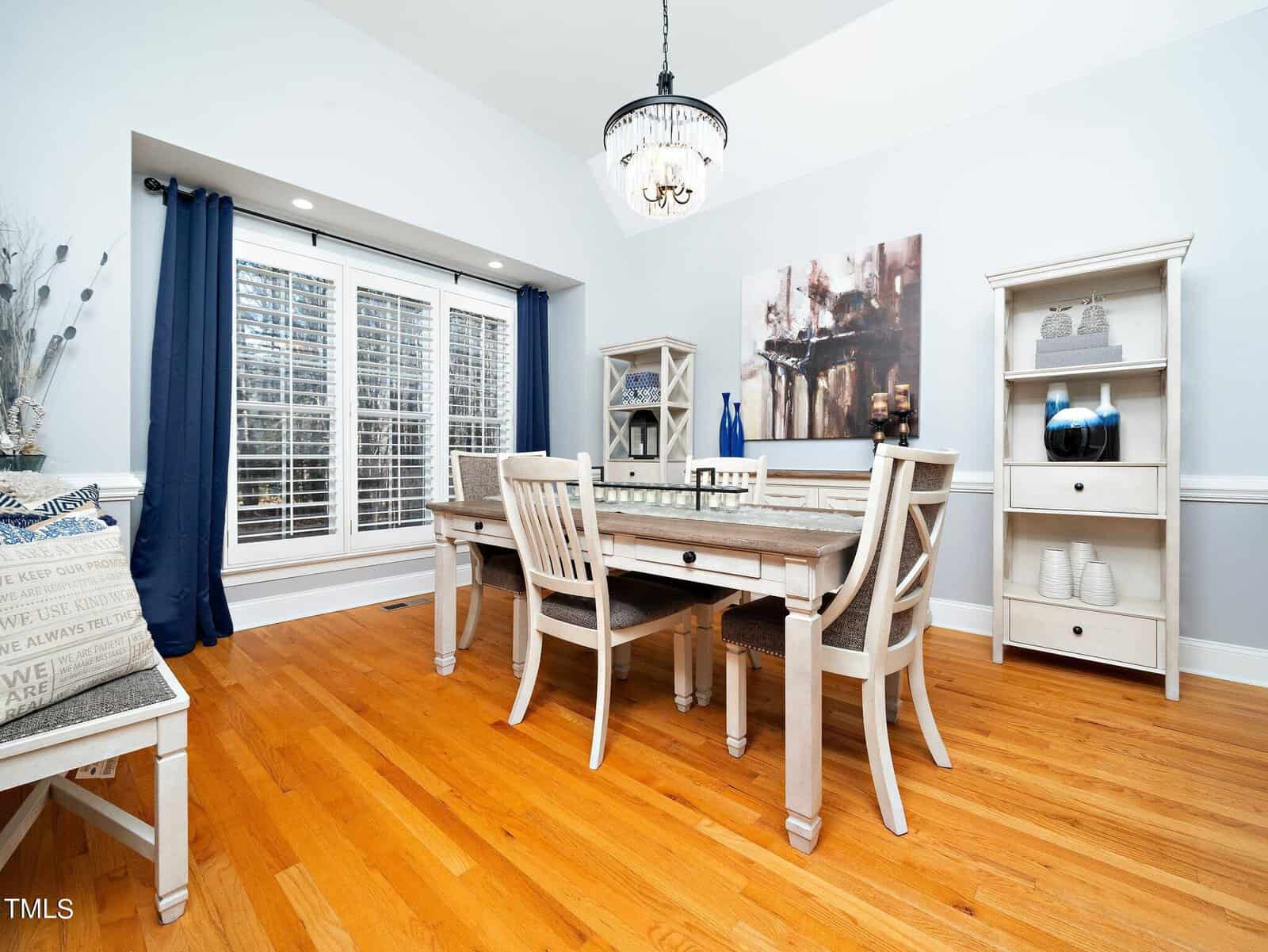 A bright dining room with a wooden floor features a rustic wooden table surrounded by six chairs. A chandelier hangs from the ceiling. The room has white and blue decor, large windows with shutters, and a painting on the wall. Shelving units flank the table.