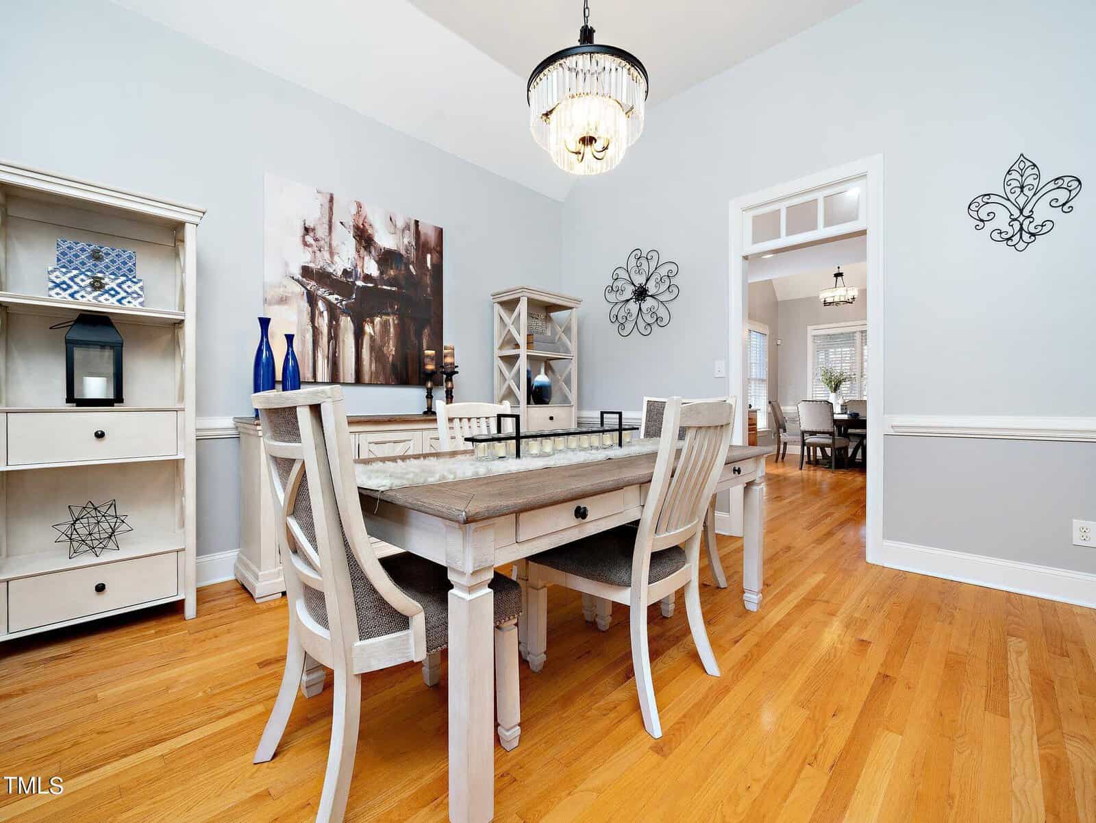 A bright dining room with a wooden table and six chairs on a hardwood floor. A chandelier hangs above the table. The room has light blue walls with white trim, decorative wall art, and shelving units displaying decor items.