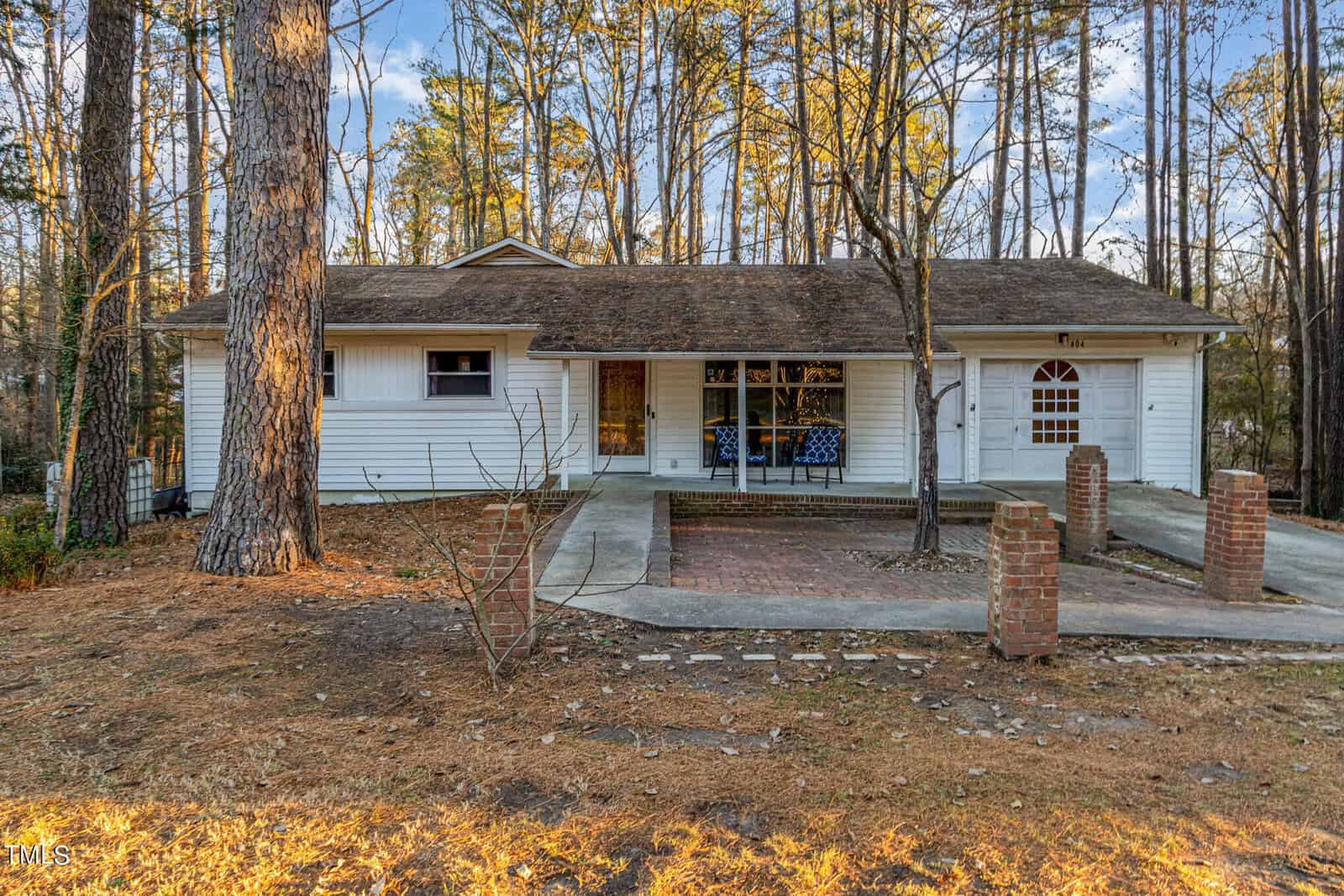 A single-story white house on 404 E D Street in Butner boasts a brown shingle roof and is surrounded by tall trees. The front features a covered porch and a brick-paved area. A curved driveway leads to a red brick entryway framed by two brick pillars.