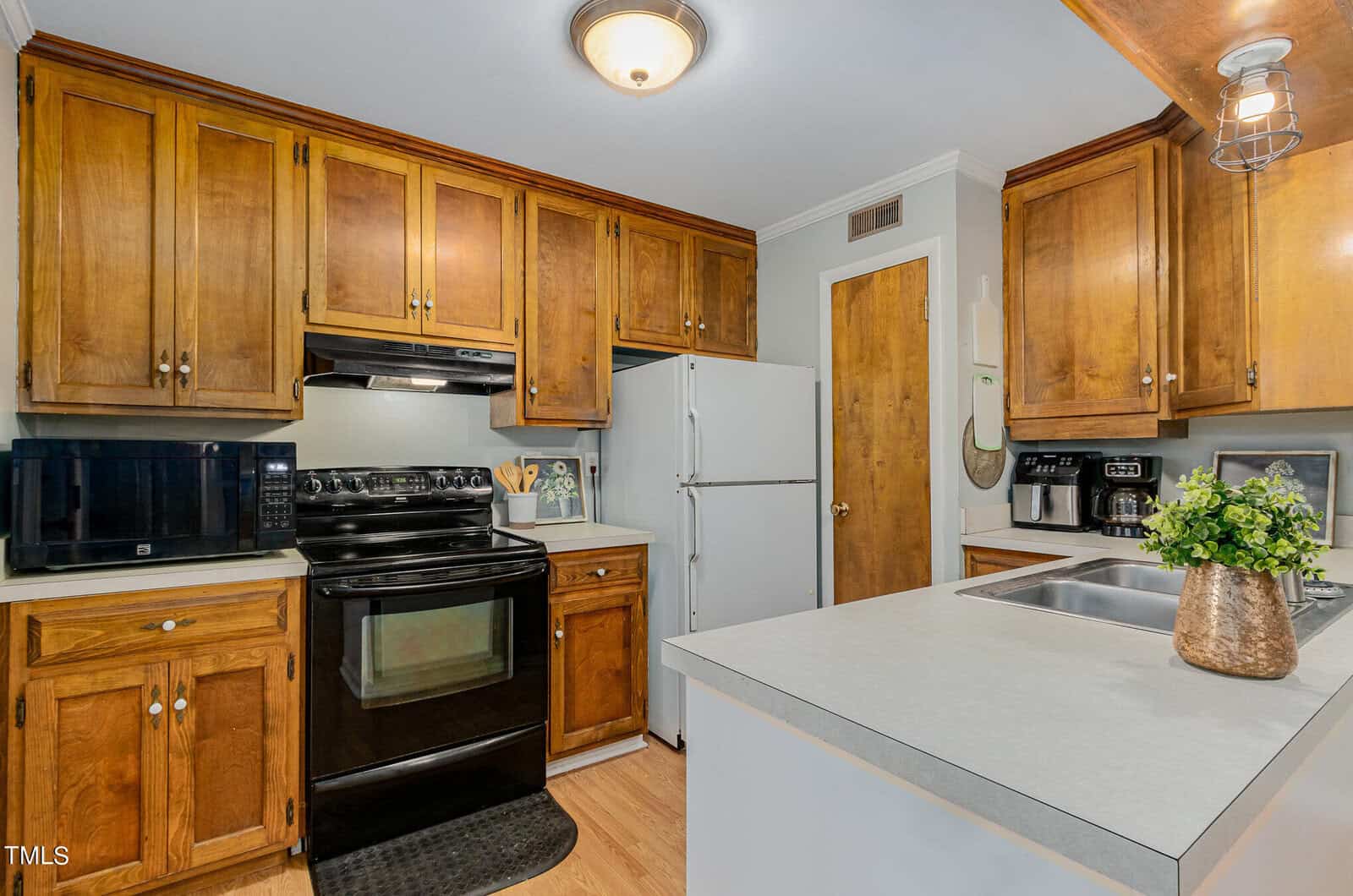 A cozy kitchen with wooden cabinets, black appliances, and a white refrigerator. A small island with a sink is centered, adorned with a potted plant. The room has overhead lighting and a microwave on the countertop.
