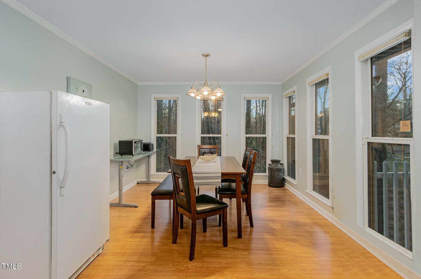 A dining room with a wooden table set for six, surrounded by chairs on a light hardwood floor. Large windows offer a view outside. A white refrigerator and a small black microwave are on the left. Warm overhead lighting creates a cozy ambiance.