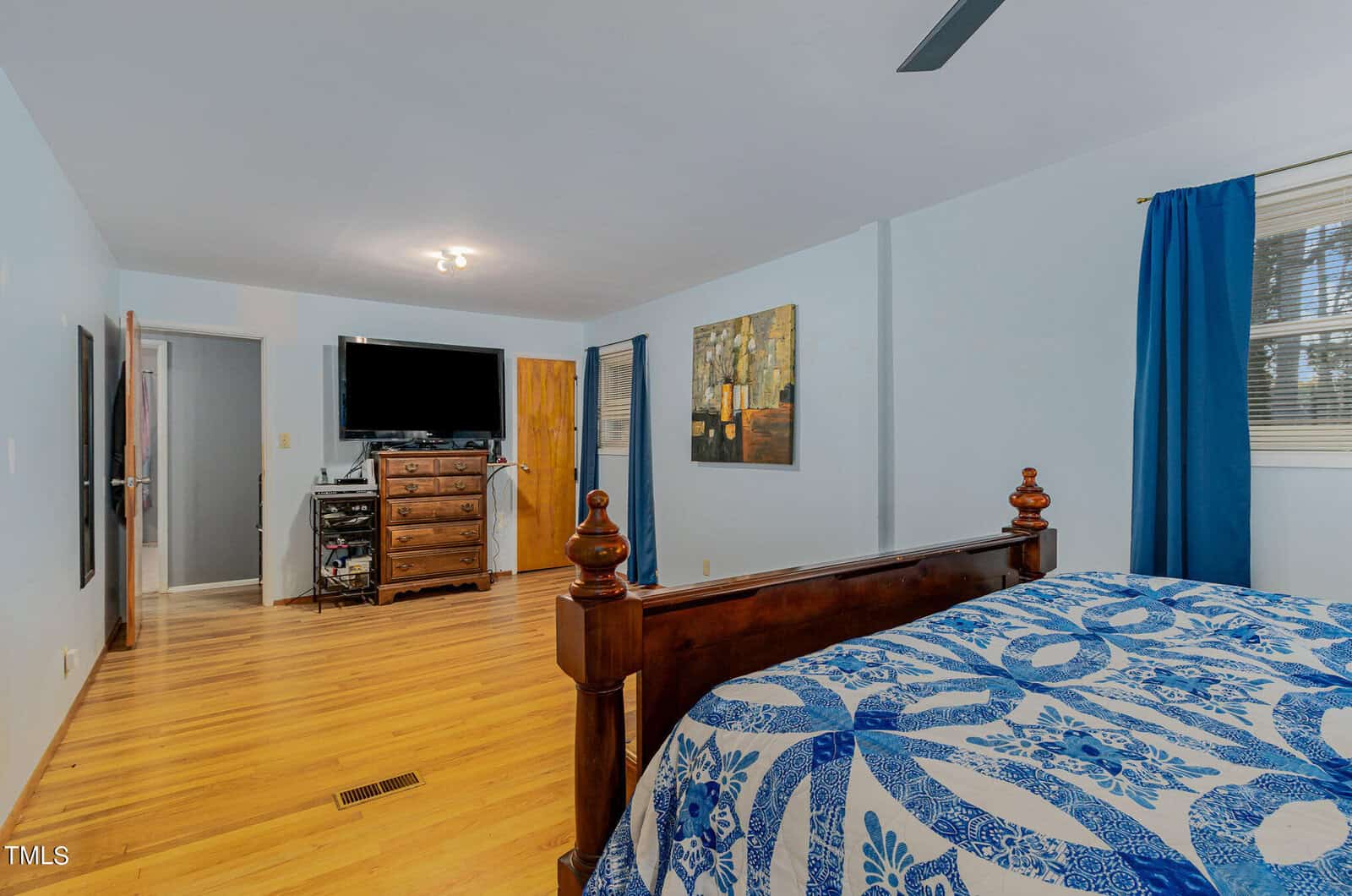 Bedroom with light blue walls, featuring a dark wooden bed with a blue and white patterned quilt. A dresser with a TV on top is against the far wall, and two windows with blue curtains let in natural light. Wood flooring adds warmth to the room.
