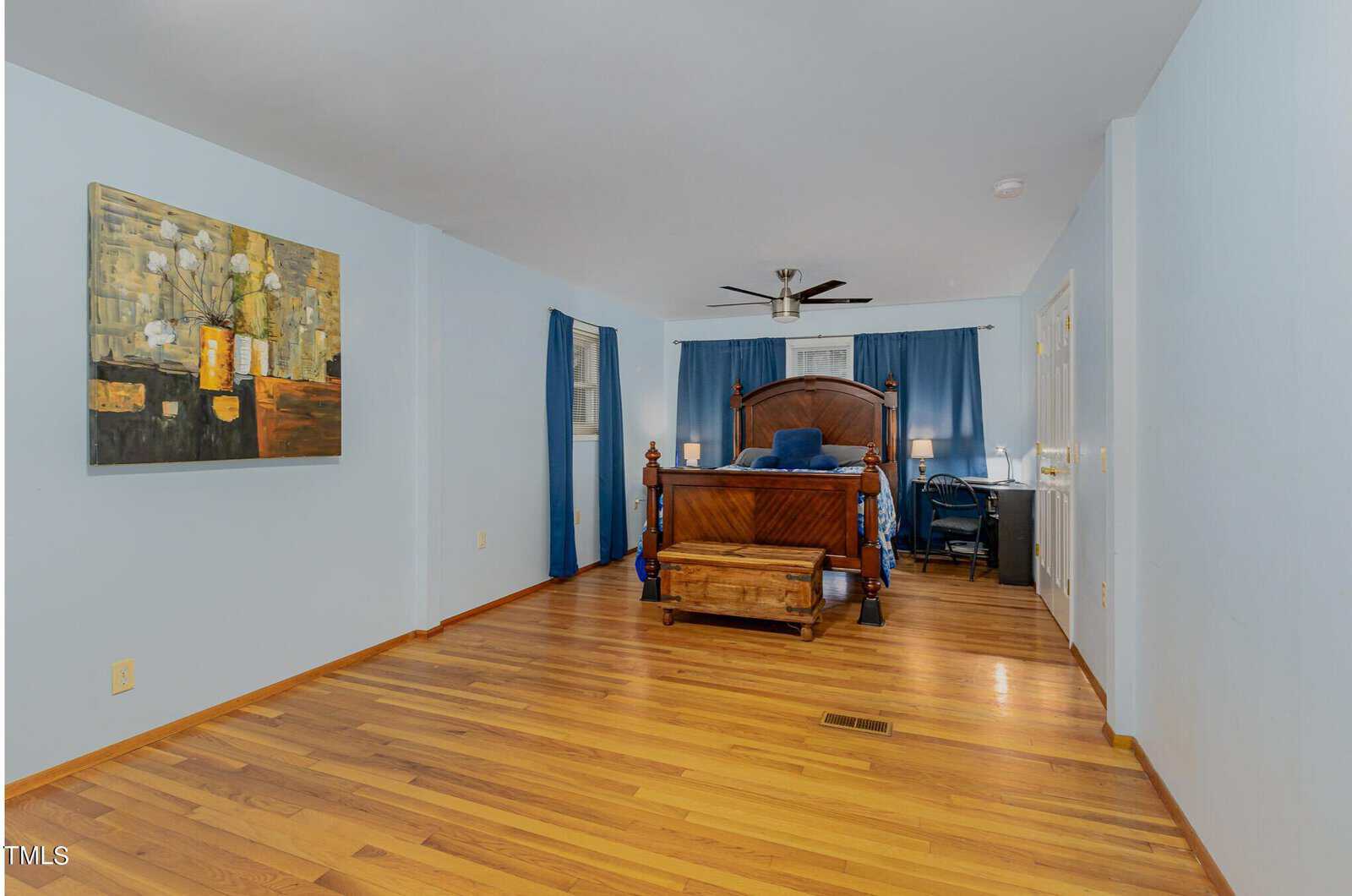 A spacious bedroom with hardwood floors and light blue walls. It features a wooden bed with blue bedding, a chest at the foot, a painting on the left wall, dark blue curtains, and a ceiling fan.