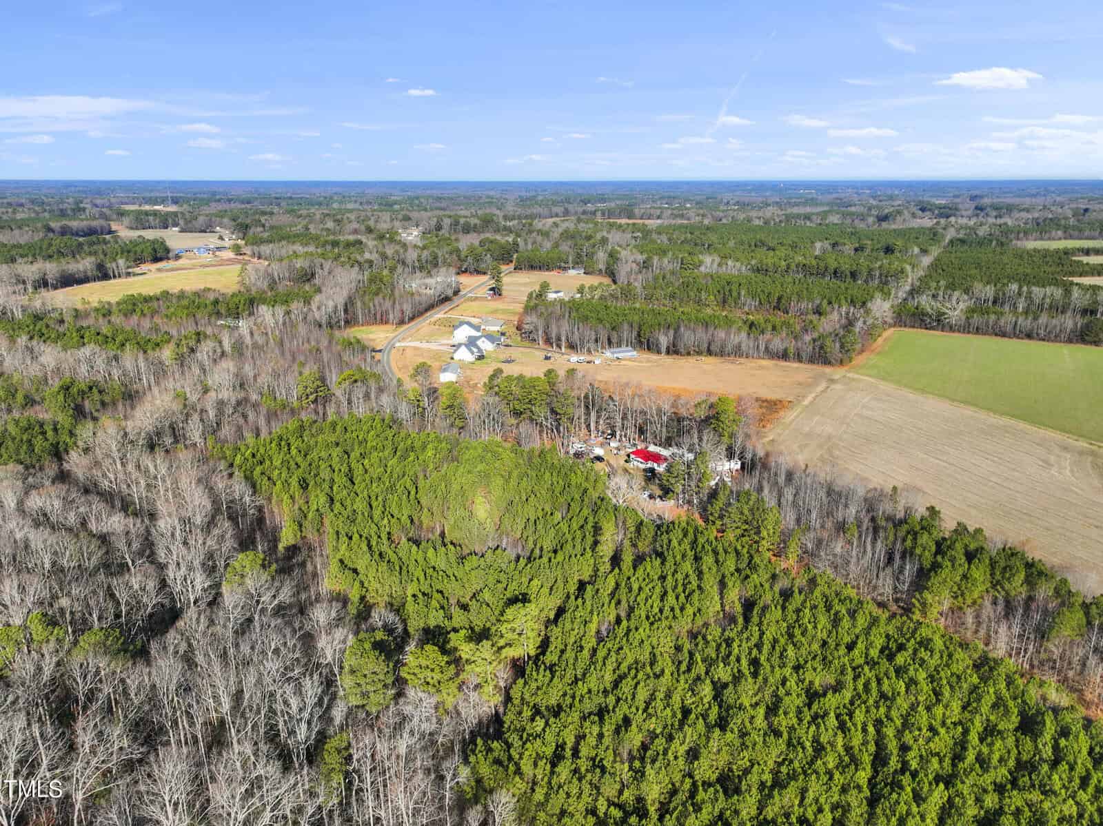 Aerial view of a rural landscape with green forests, open fields, and scattered buildings under a blue sky. A cluster of farm structures and a red-roofed building are visible near the center of the image.