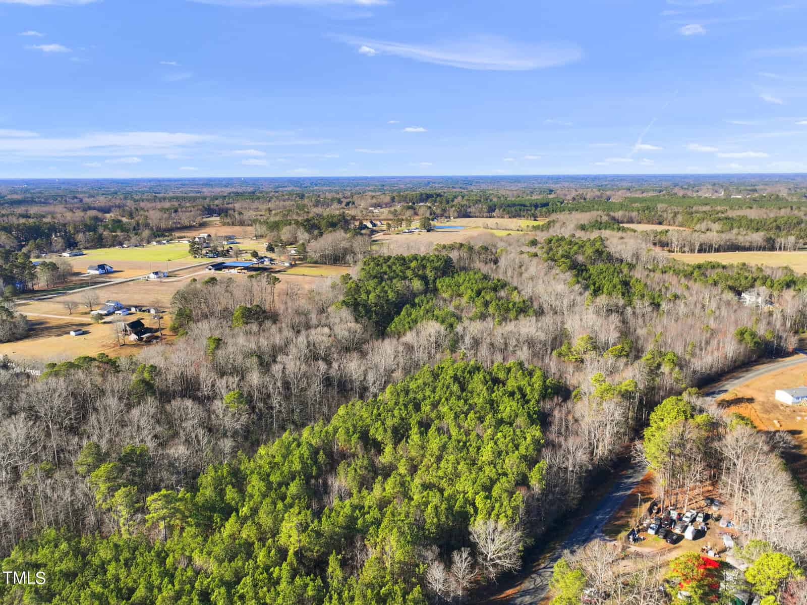 Aerial view of a rural landscape with dense green forests, scattered houses, and open fields under a clear blue sky. A small dirt road and parked vehicles are visible in the lower right corner.