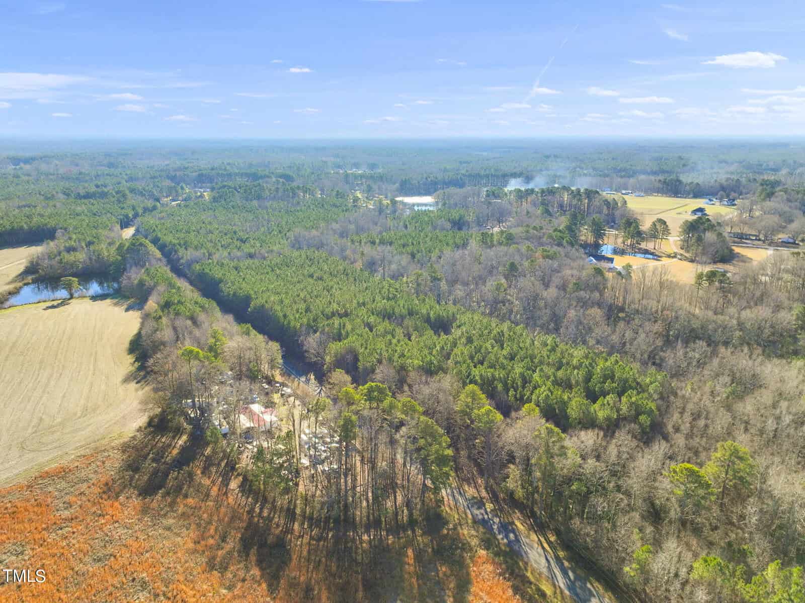 Aerial view of a rural landscape with dense forests, open fields, scattered houses, and winding roads under a clear blue sky.