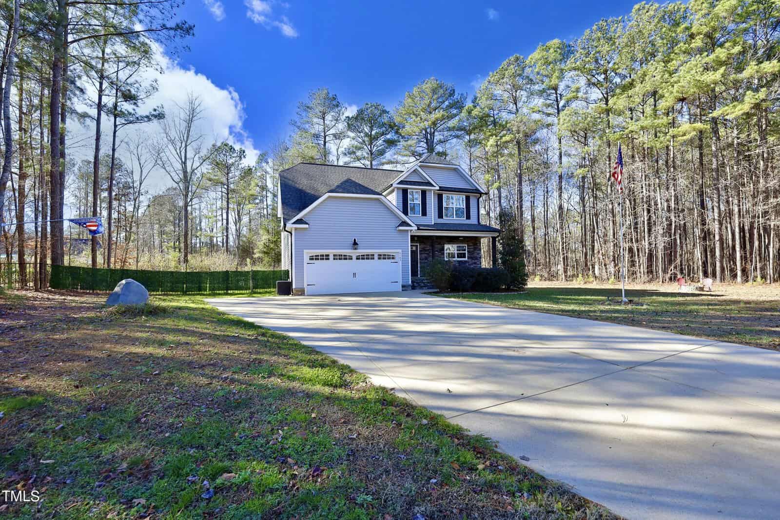 A two-story house with a white garage sits at the end of a long driveway surrounded by a lush forest. A flagpole is on the right, and a bright blue sky with a few clouds is overhead. The yard is spacious and partially shaded.