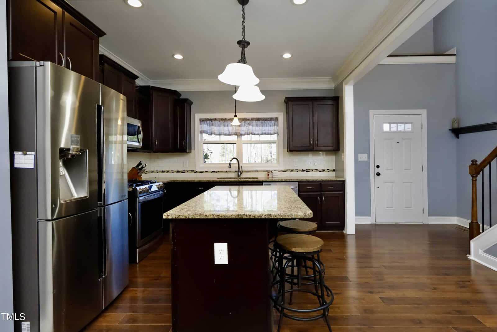 Modern kitchen with dark wood cabinets, stainless steel appliances, and a large granite island with bar stools. A hanging light fixture illuminates the space. A white door and staircase are visible to the right.