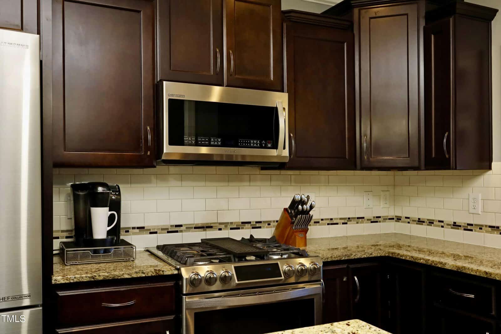 A modern kitchen with dark wood cabinets, a stainless steel oven and microwave, granite countertops, a knife block, and a coffee maker. The backsplash features white subway tiles with decorative accents.