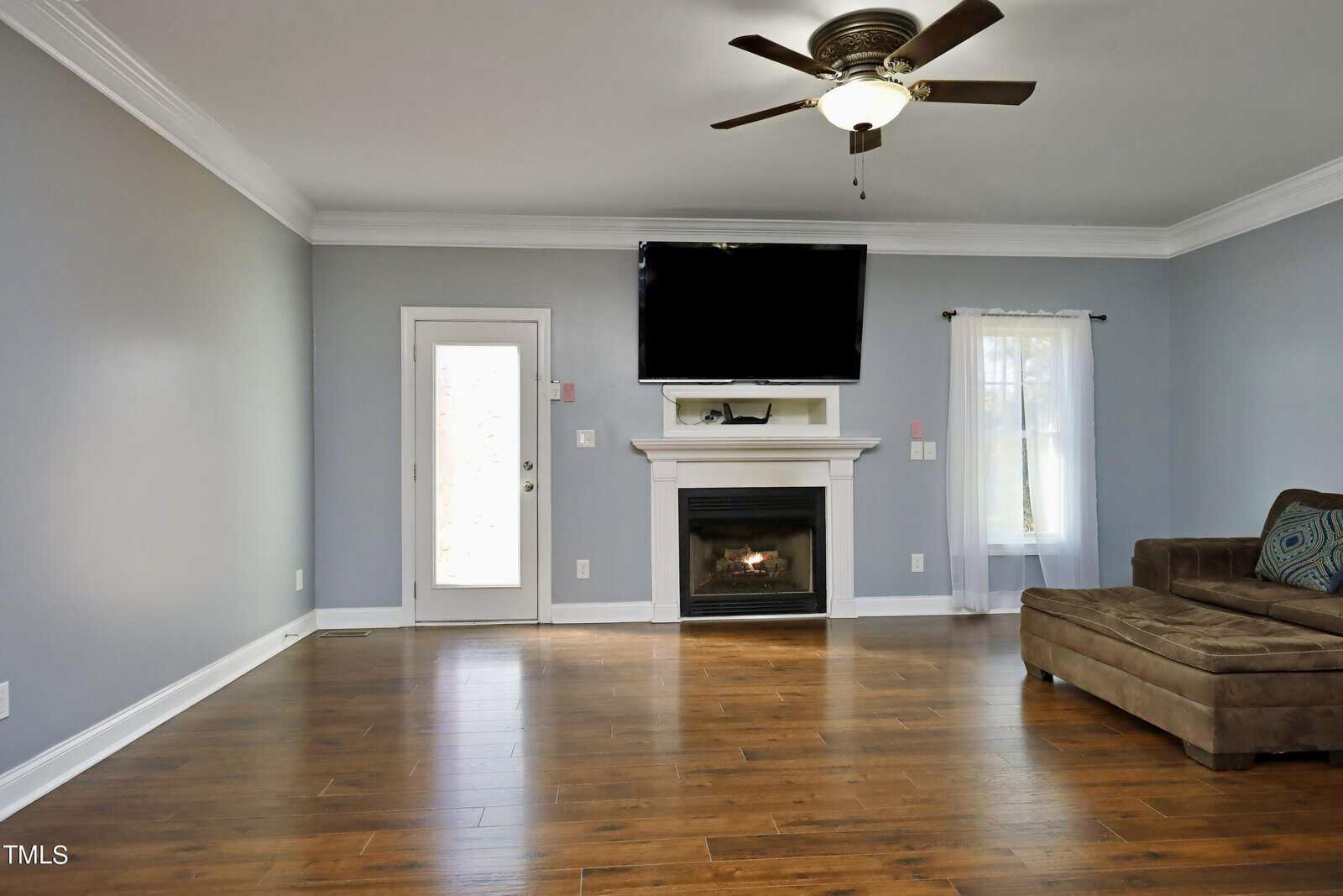 A spacious living room with light gray walls and hardwood flooring. There's a ceiling fan and a wall-mounted TV above a fireplace. A brown sectional sofa sits on the right, and a glass door is on the left with natural light streaming in.