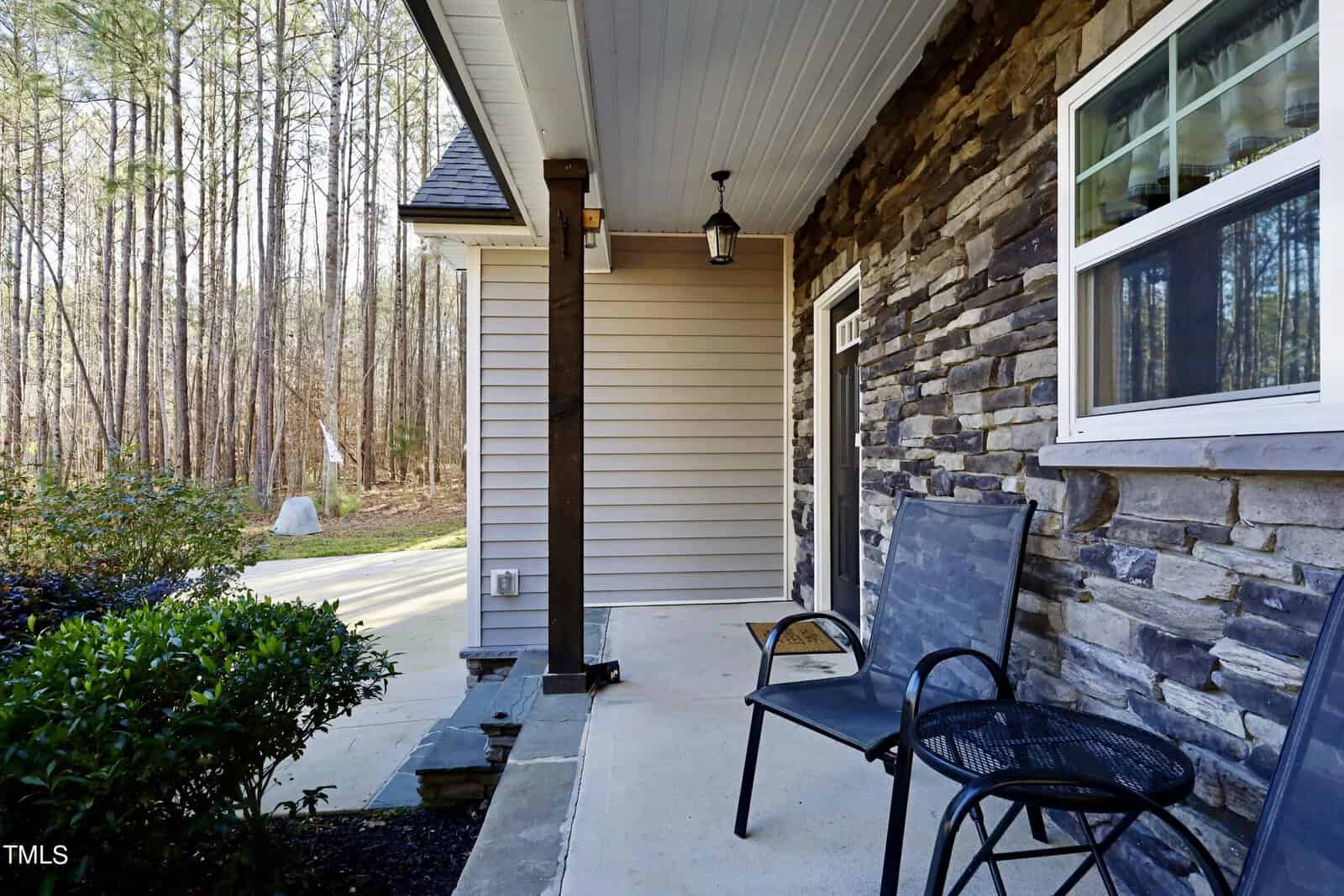 A cozy porch with two metal chairs and a small round table. The house exterior features stone and siding. A pathway leads to a wooded area in the background. A hanging light fixture adds charm to the porch.