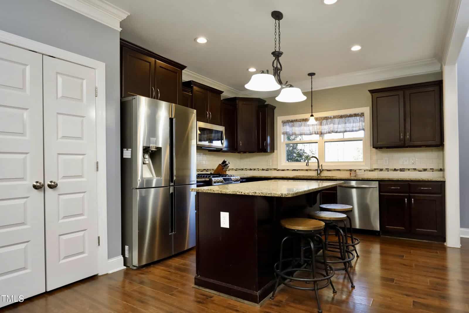 Modern kitchen with dark wooden cabinets, granite countertops, stainless steel appliances, and a central island with three round stools. There's a double-door pantry and hanging pendant lights, all set on a polished wooden floor.