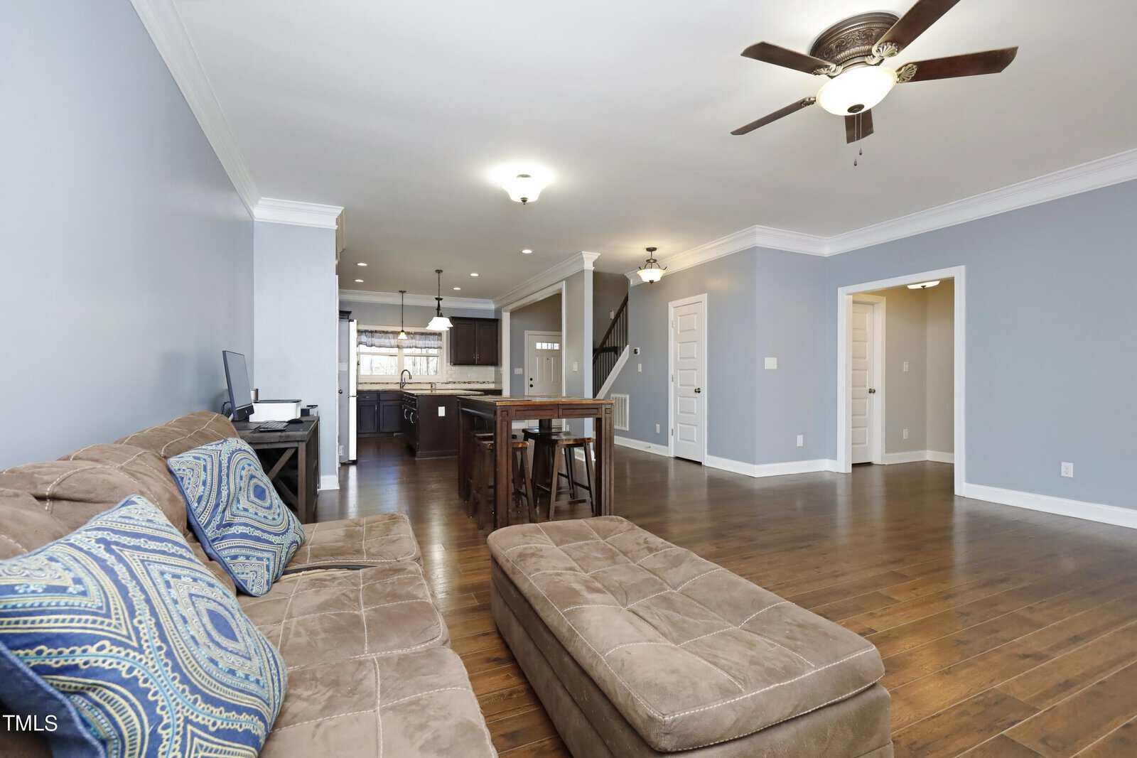 A spacious living room with light blue walls, a brown sectional sofa, and matching ottoman. The room features dark wooden floors and a ceiling fan. In the background, there is a kitchen with a modern island and bar stools.