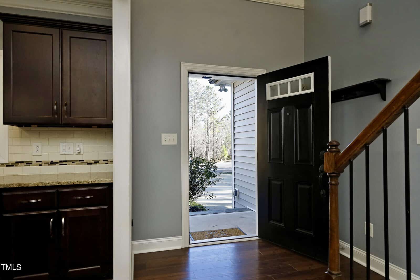 A modern interior featuring an open black front door with a small window, leading outside. On the left, there's a kitchen corner with dark wood cabinets and a granite countertop. Wooden stairs with a railing ascend on the right.
