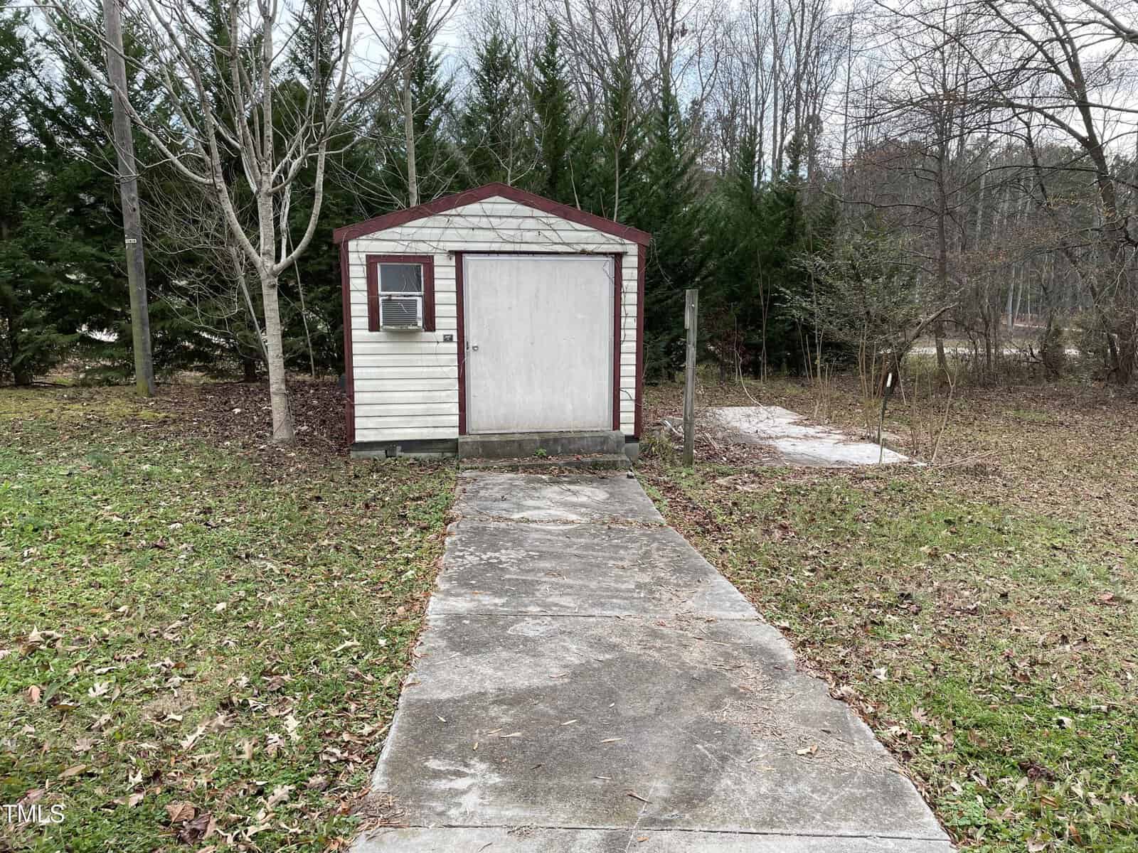 A small, white shed with a red door and window stands at the end of a concrete pathway surrounded by scattered leaves and patches of grass, with trees and a hint of snow in the background.