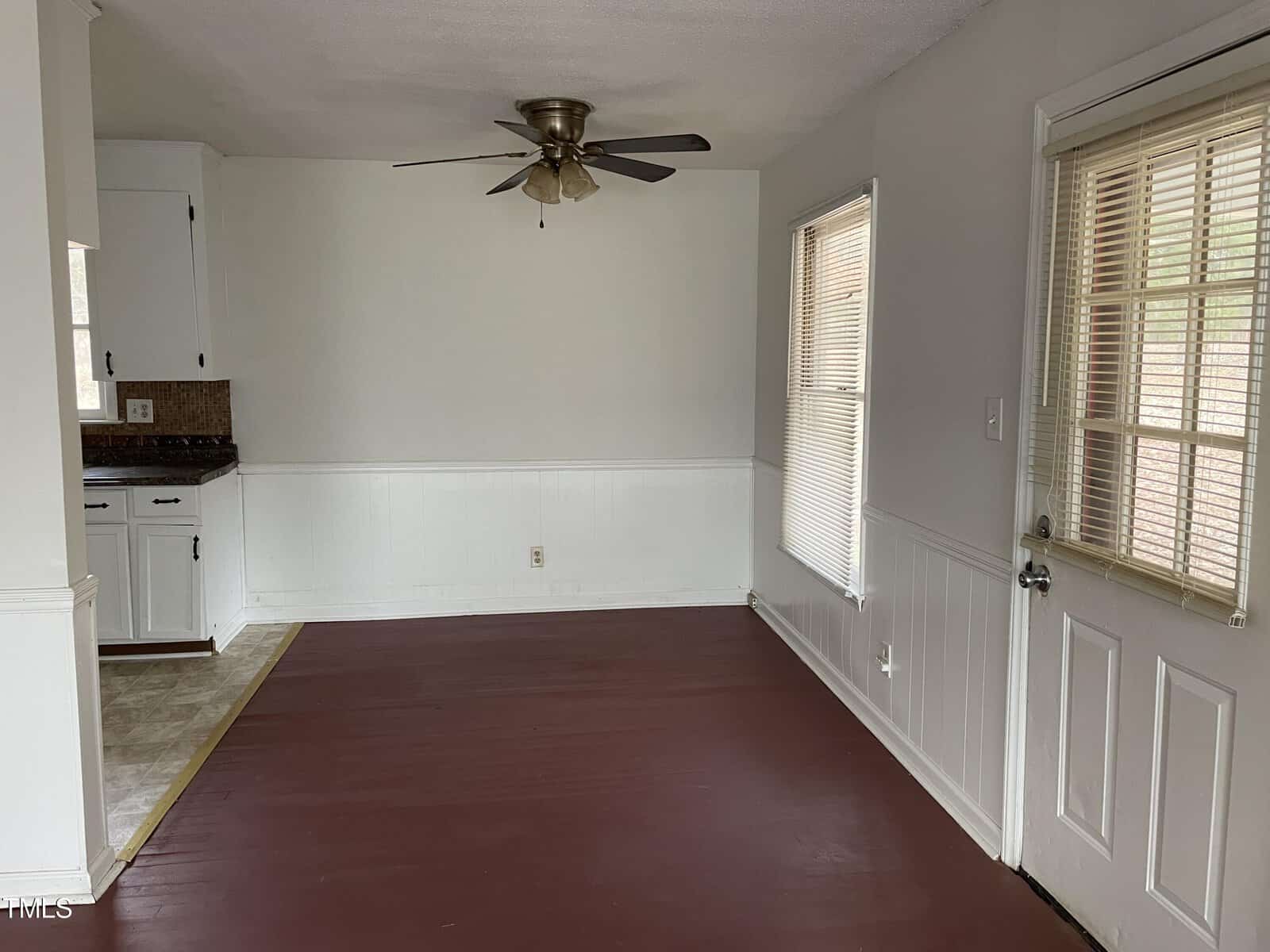 An empty room featuring white walls with a chair rail, a maroon floor, two windows with blinds, and a ceiling fan. a section of kitchen cabinets and countertops is visible in the back left corner.