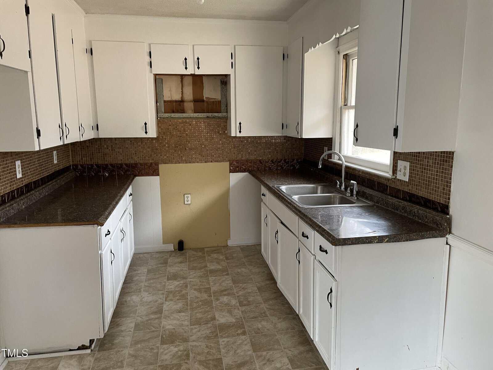A dated kitchen interior featuring white cabinetry, brown tiled countertops, and a mosaic backsplash. the room includes a double sink beneath a window and tiled flooring.