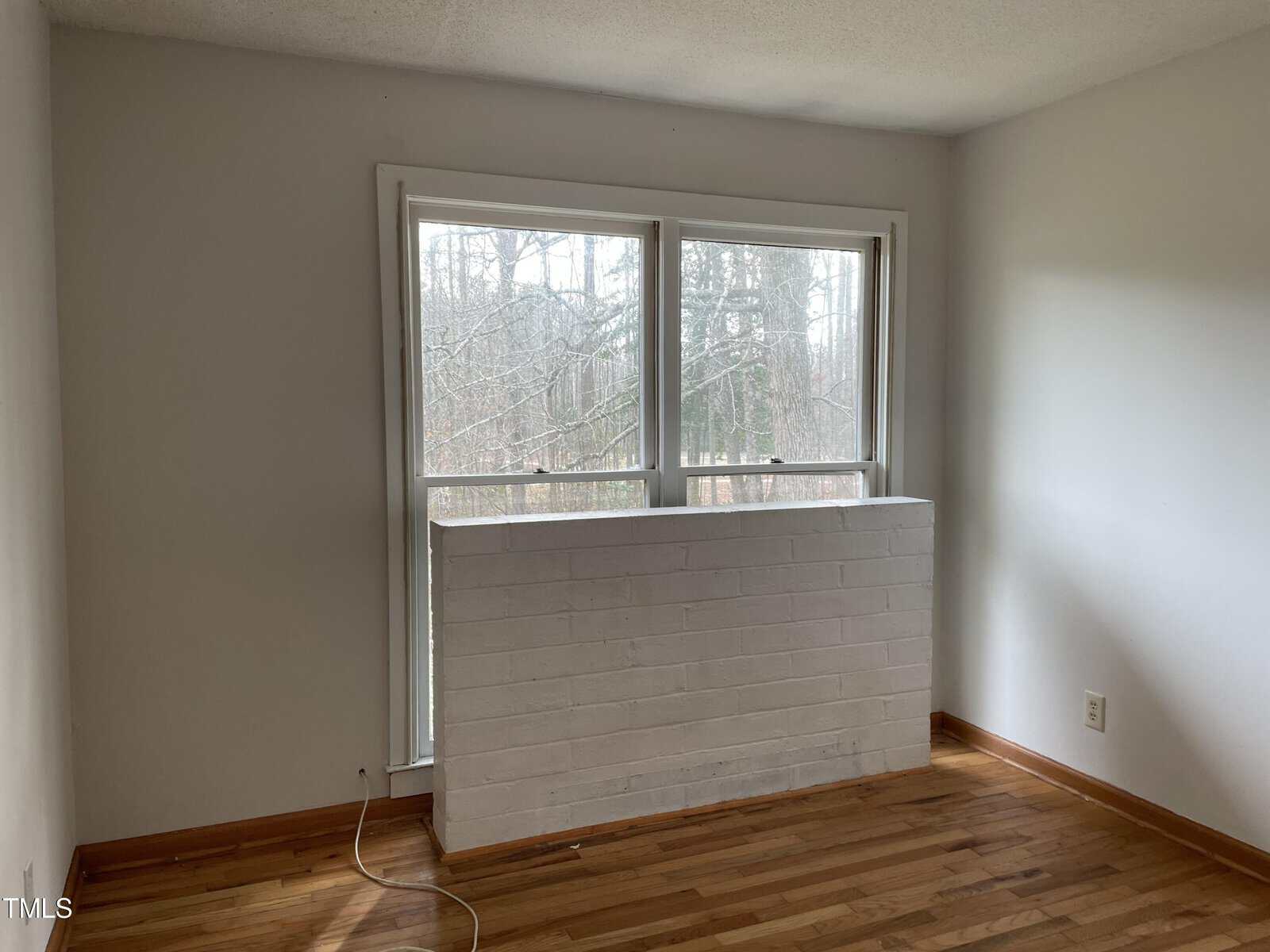 A room with bare wooden floors featuring a large window with white framing. the window overlooks leafless trees and reveals a white-painted half-brick wall beneath.