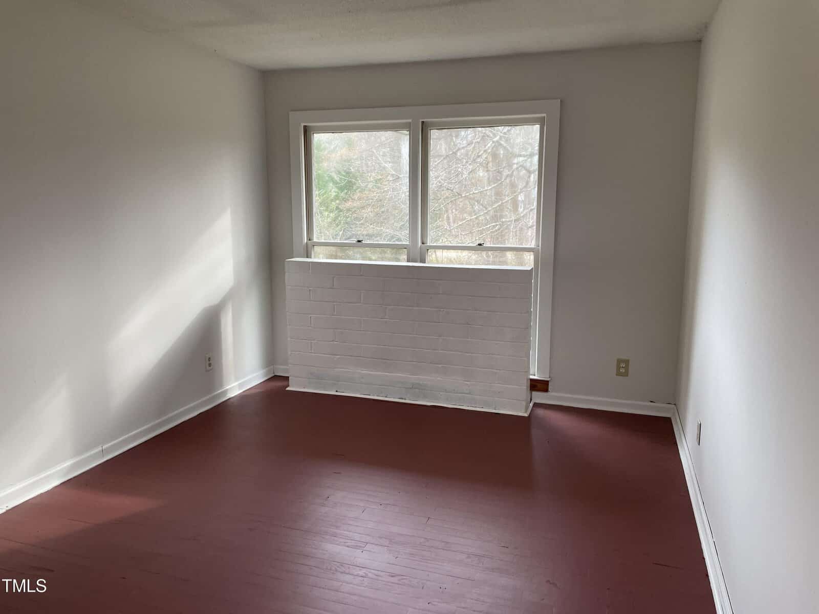 An empty room with a white brick partition and a large window that looks out to trees. the floor is covered with red hardwood. natural light illuminates the space.