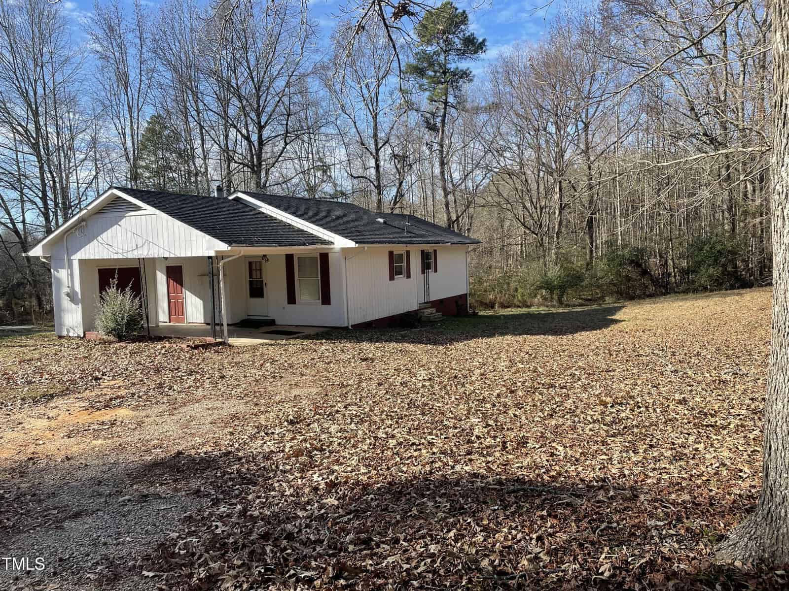 A modest white house with a red door and brown roofing, situated in a wooded area with scattered leaves on the ground and bare trees around. there is a driveway on the left side of the house.