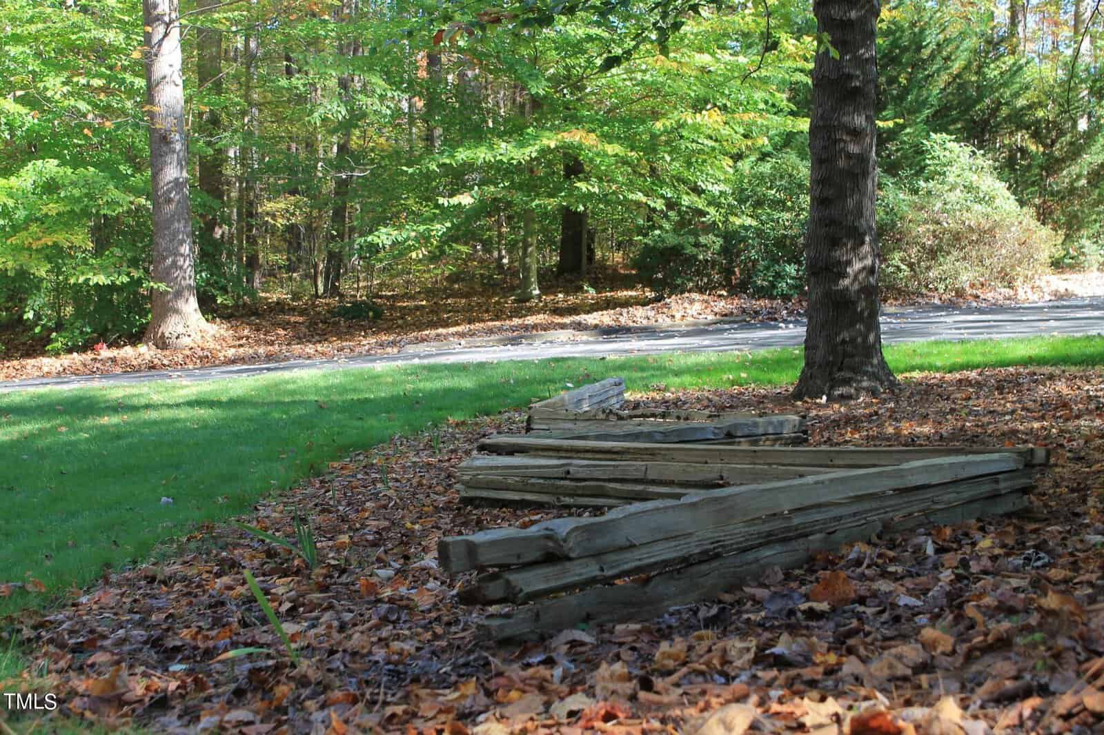 A stack of weathered wooden planks lying on a bed of autumn leaves by the side of a road with trees in the background.